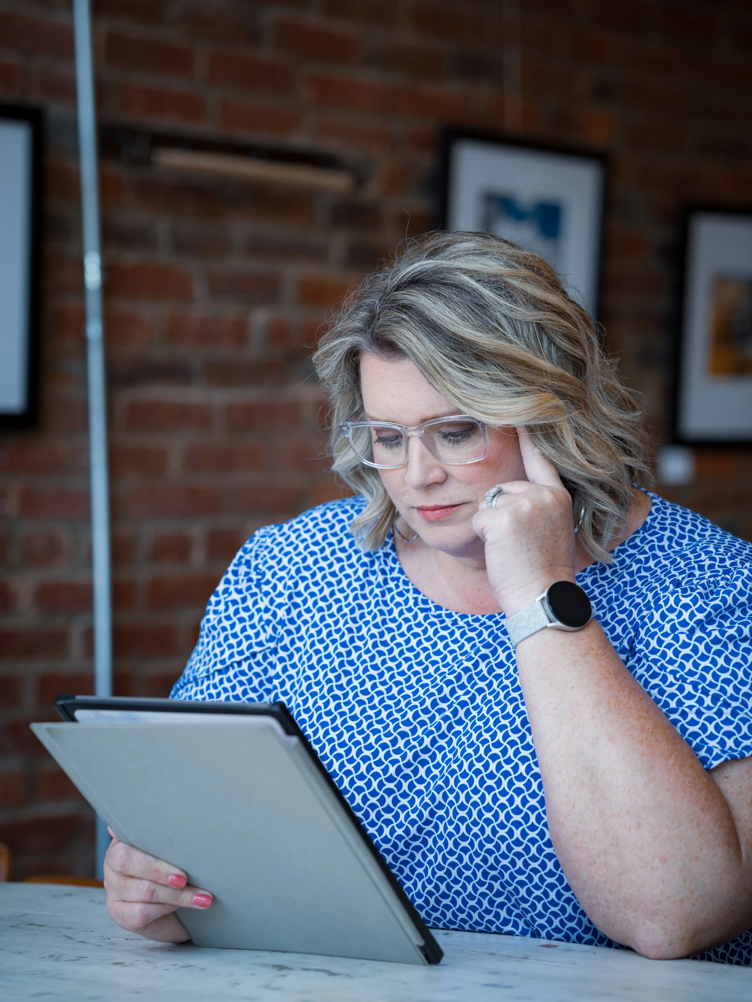 A woman with blond hair and wearing glasses is sitting at a table, reading a tablet. She is dressed in a blue patterned shirt and has a smartwatch on her wrist. Behind her, there are framed pictures on a brick wall.
