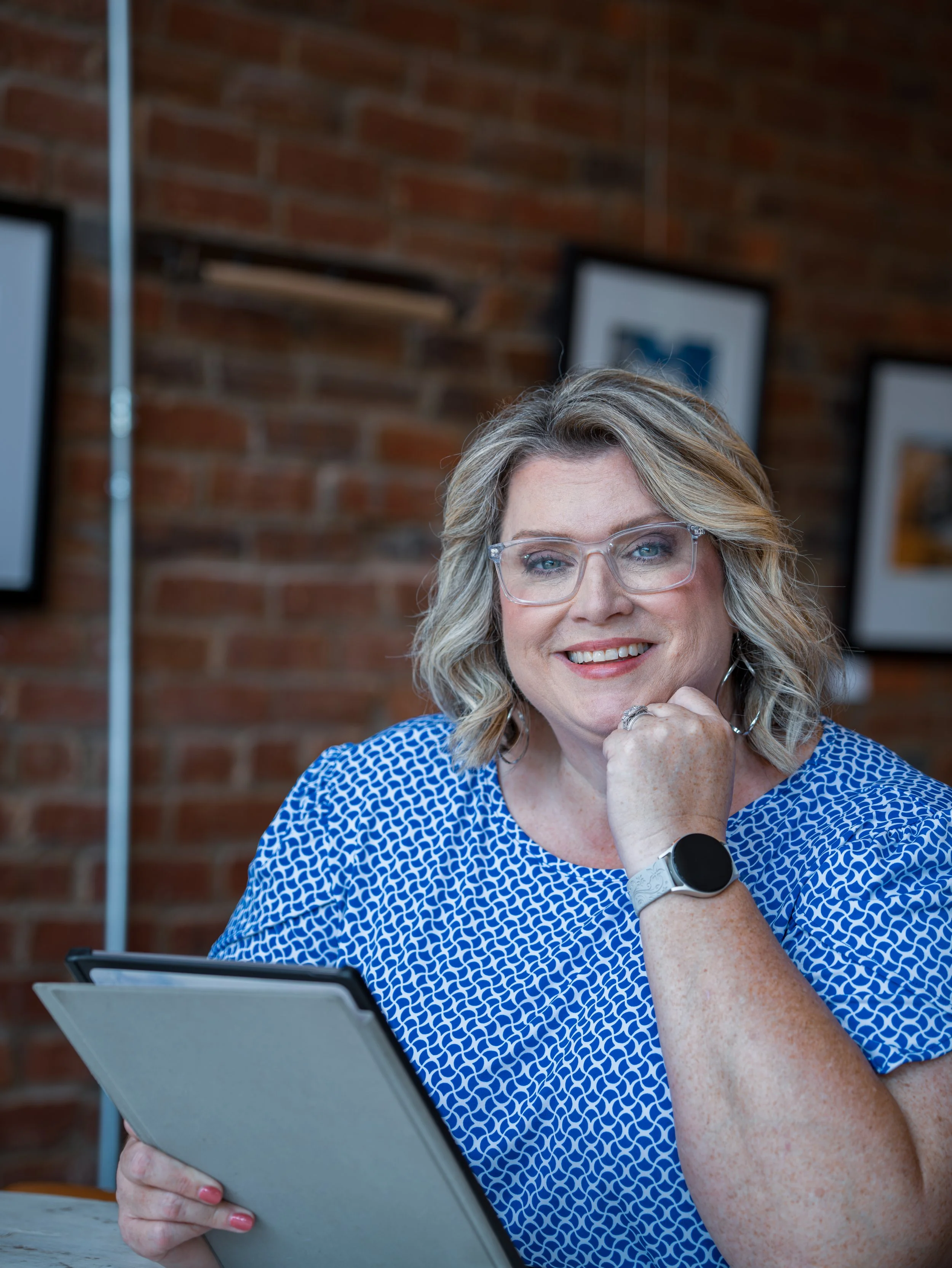 A woman with blonde hair and glasses smiling while looking at a tablet in a room with brick walls and framed pictures.