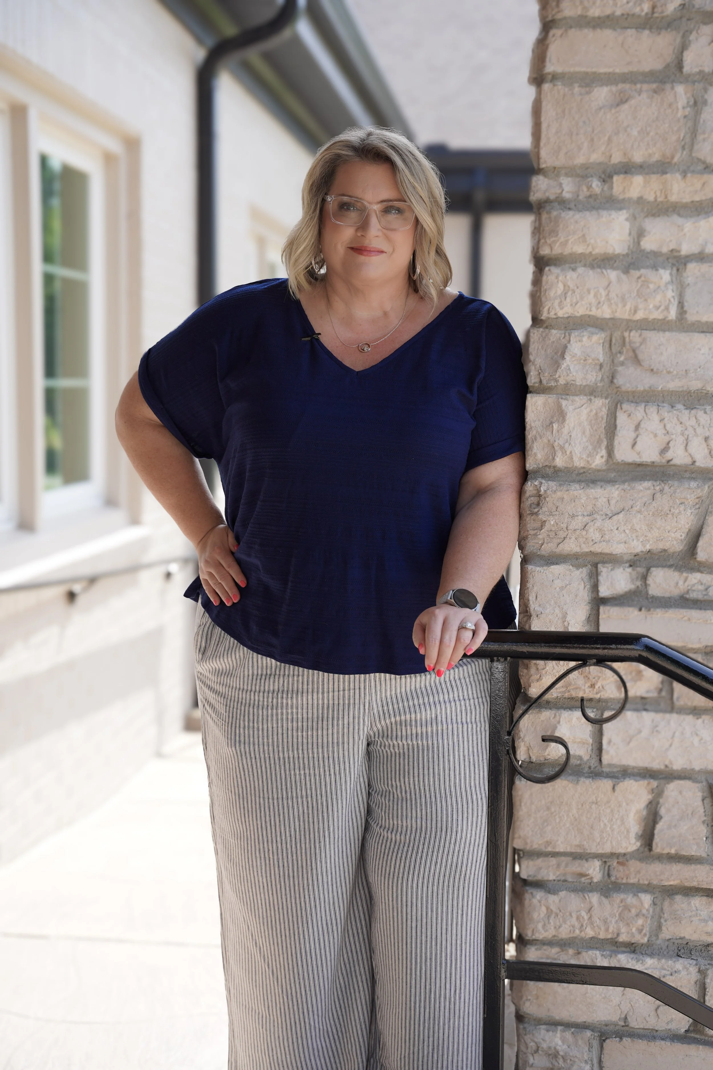 A woman with blonde hair, glasses, and pink lipstick leaning on a railing outdoors, wearing a navy blue top, striped pants, and jewelry, next to a brick wall on a sunny day.