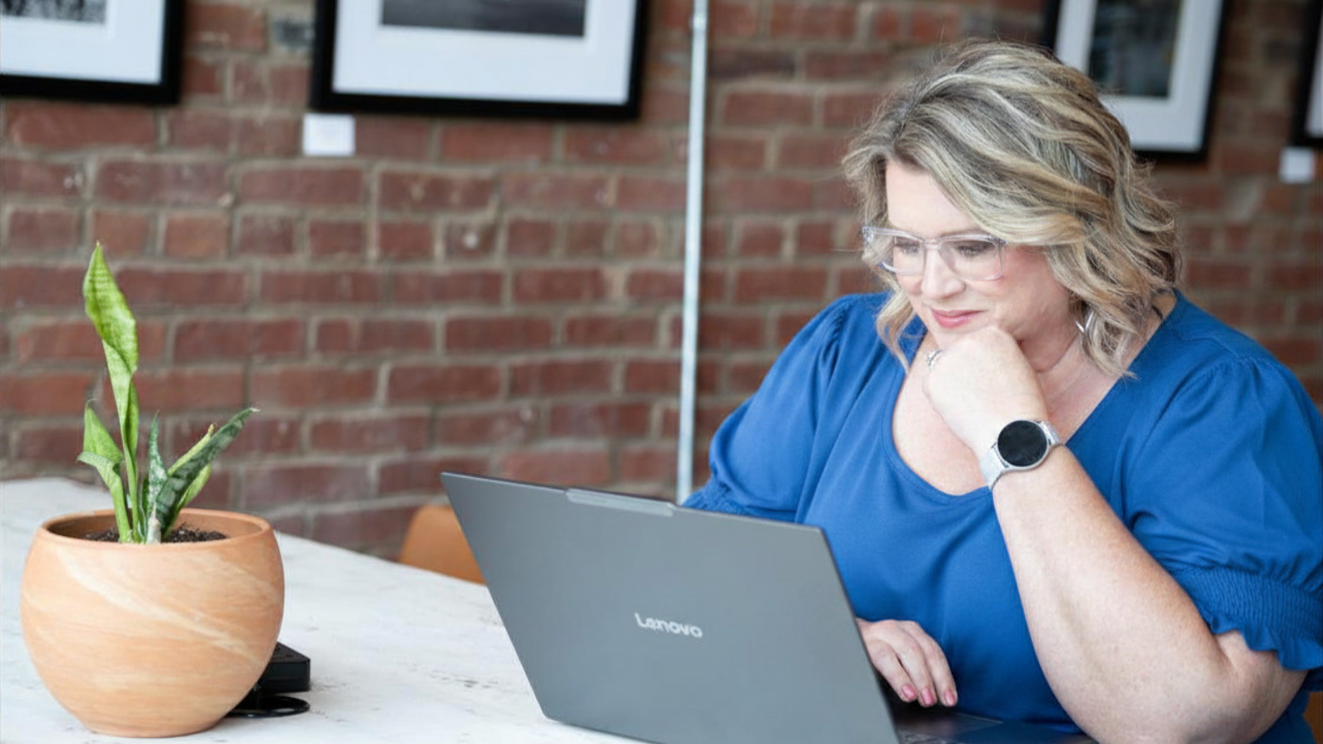 A woman with wavy blonde hair, wearing safety glasses and a blue shirt, is sitting at a table, looking at a silver Lenovo laptop. There is a potted green plant in a terracotta pot on the table, and a brick wall with framed pictures in the background.