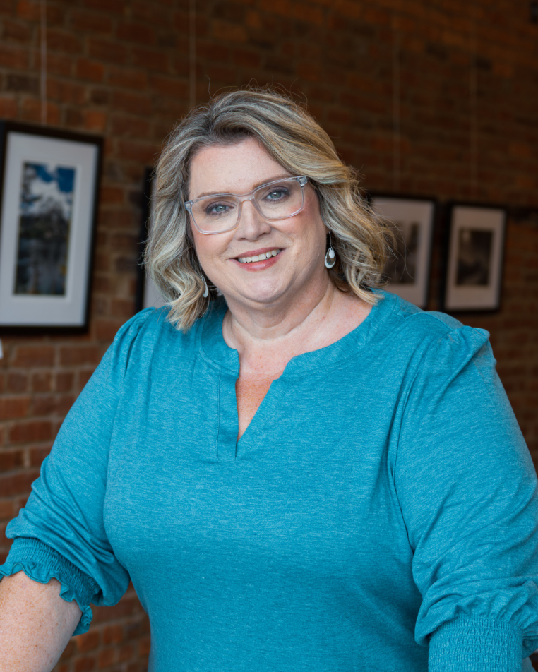 A smiling woman with blonde wavy hair, wearing glasses and a teal top, standing in front of a brick wall with framed photographs.