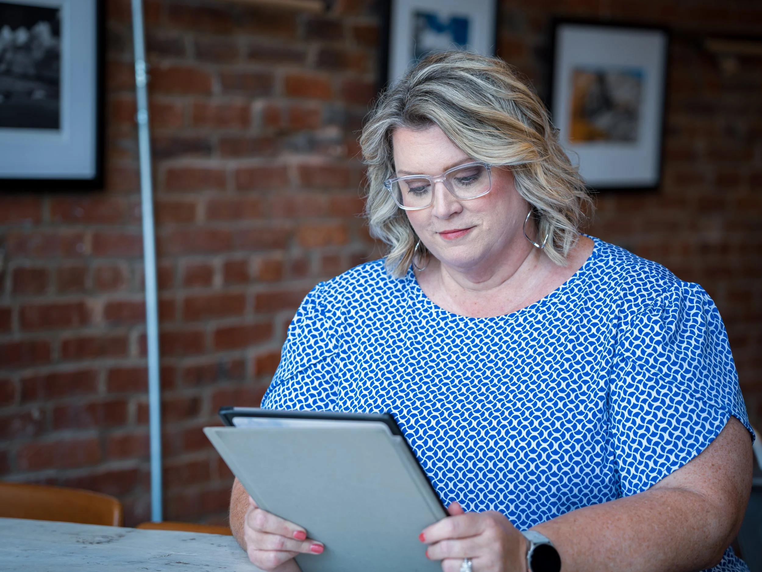 A woman with blonde hair wearing glasses and a blue patterned shirt is sitting at a table, looking down at a tablet device she is holding in her hands. There are framed pictures on a brick wall in the background.