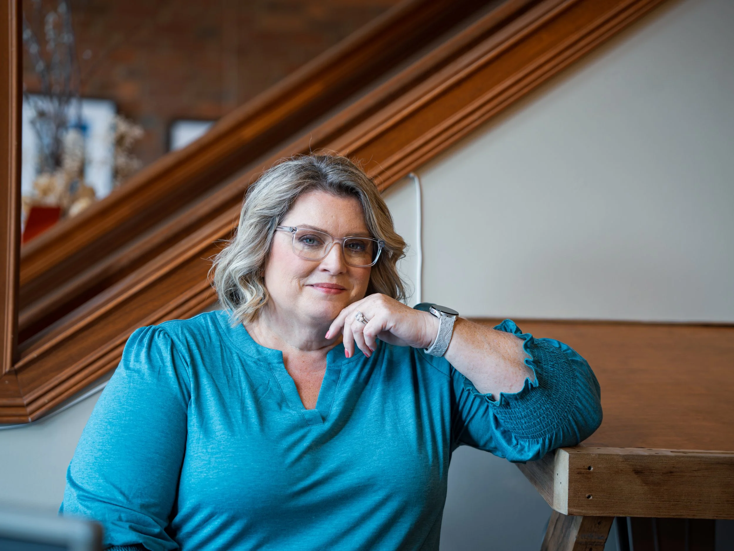 A woman with glasses and shoulder-length wavy hair, wearing a teal long-sleeve shirt, leaning on a wooden railing indoors, with a background of wooden paneling and a blurred window.