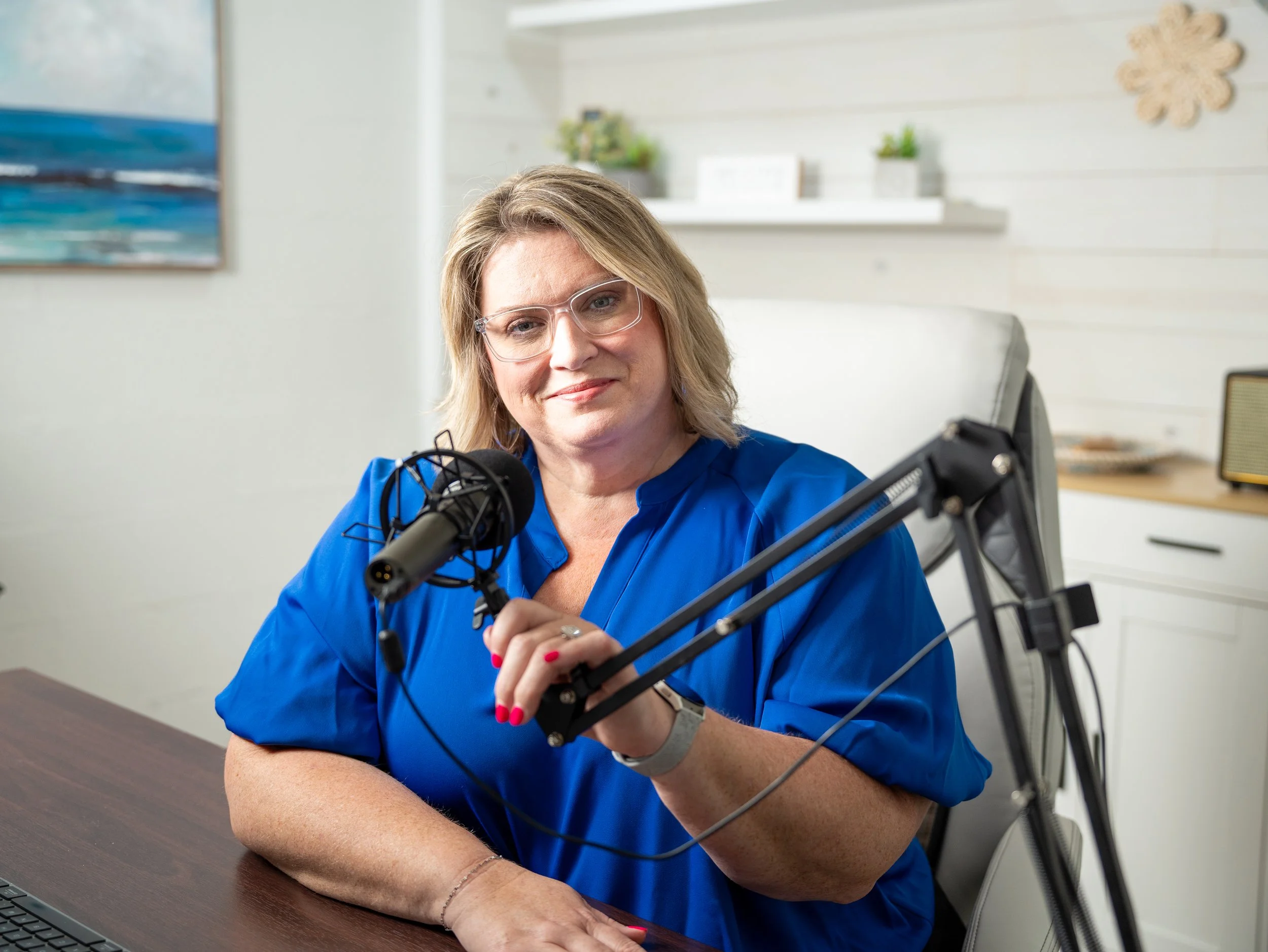 A woman with blonde hair and glasses wearing a blue shirt, sitting at a desk with a professional microphone.