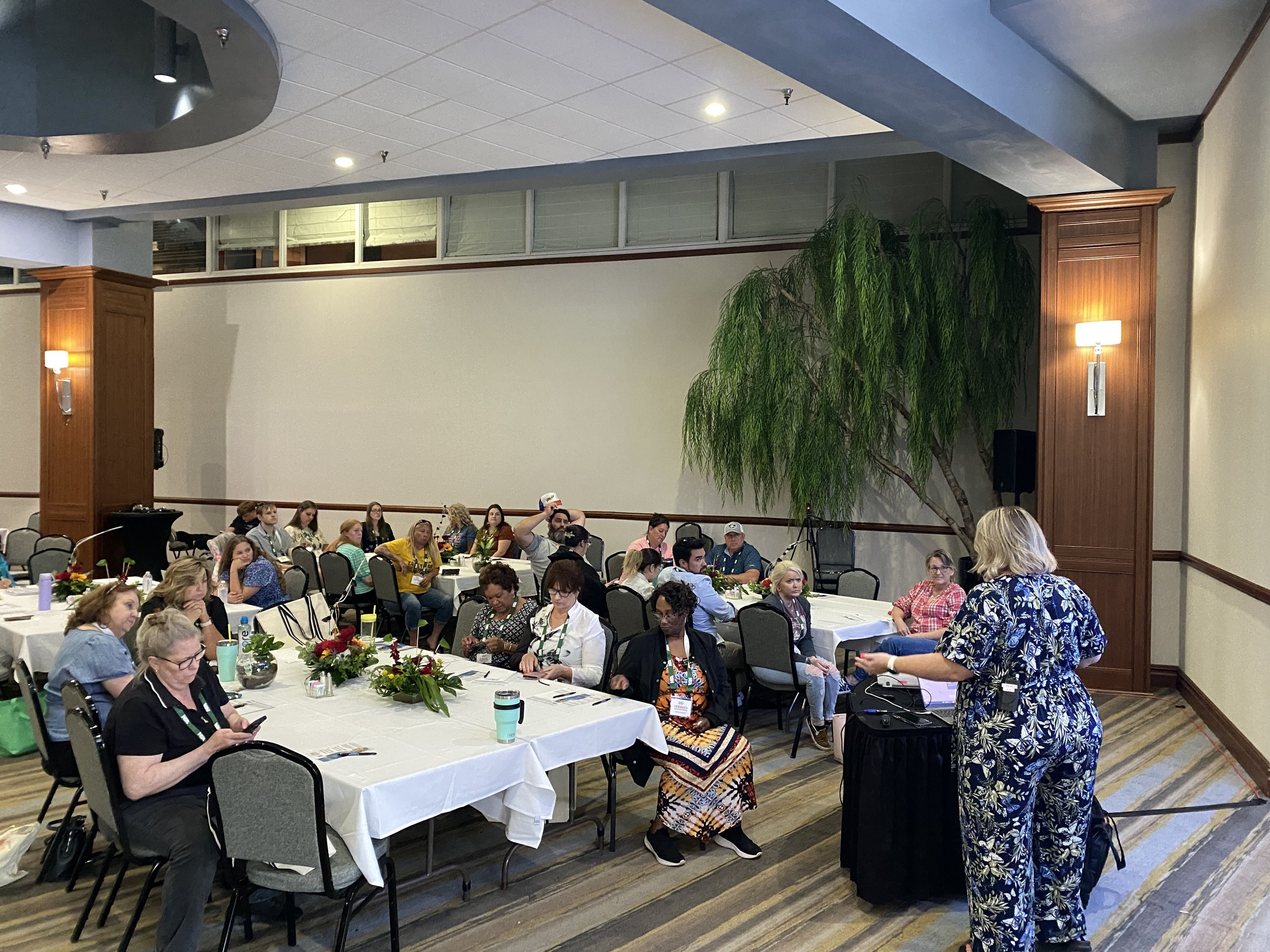 Conference room with round tables, attendees listening to a speaker at the front, and greenery in the corner. The room has a high ceiling with recessed lighting and wooden wall panels.