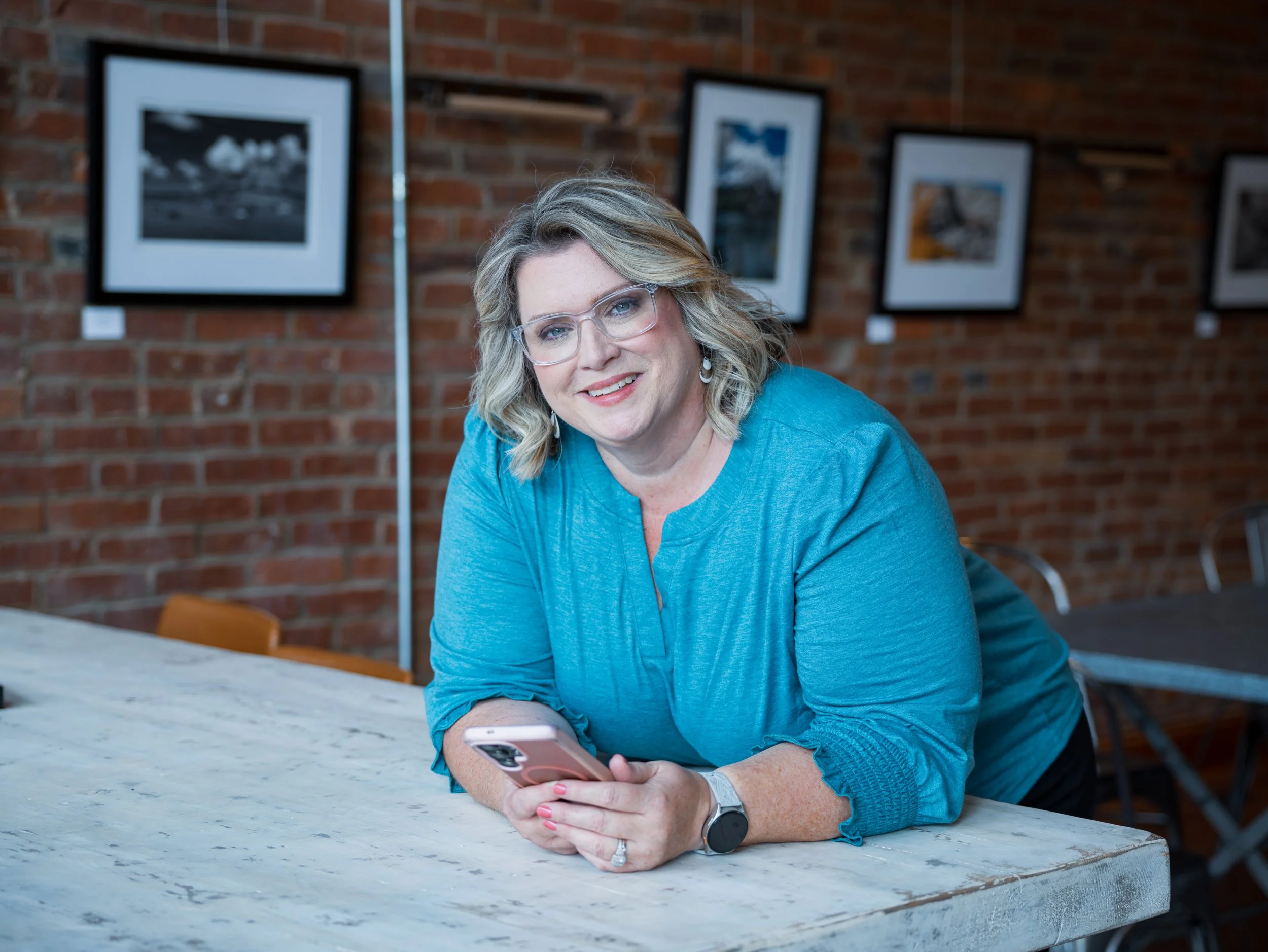 A woman with blonde hair, wearing glasses and a blue top, sitting at a marble table in a room with a brick wall in the background. She is holding a smartphone in her right hand and smiling at the camera.