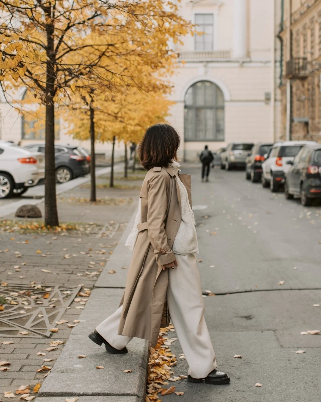 Woman in stylish white pantsuit walks across a tree lined street in autumn