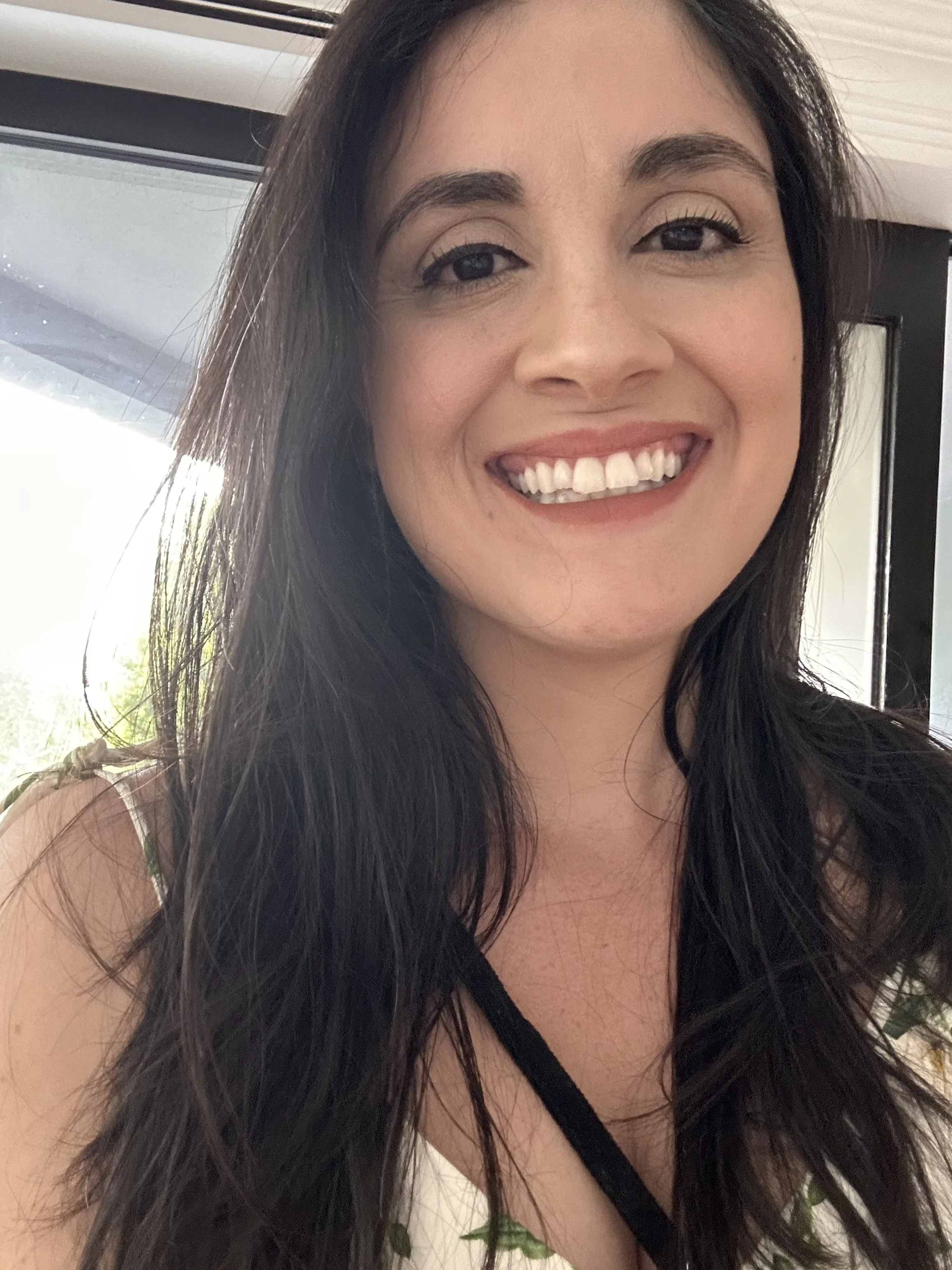 A smiling woman with long dark hair and light makeup, wearing a sleeveless top with leaf patterns, standing indoors with a window and a door in the background.