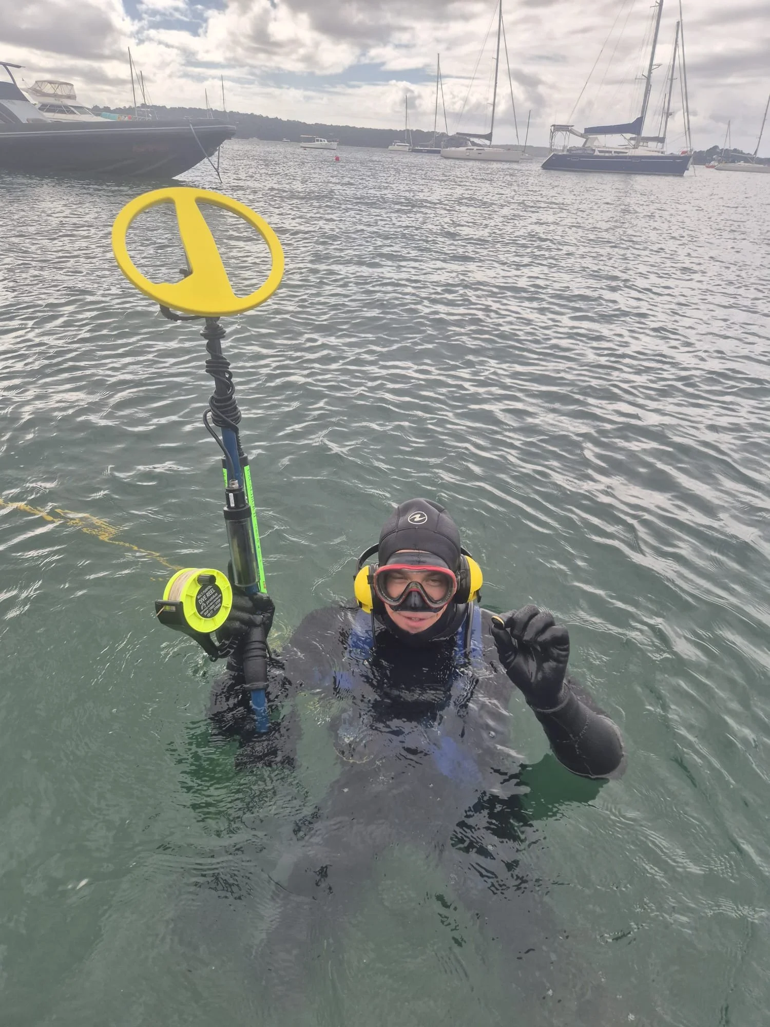 Person in scuba diving gear and wetsuit emerging from water, holding a metal detector after conducting search and recovery operations.