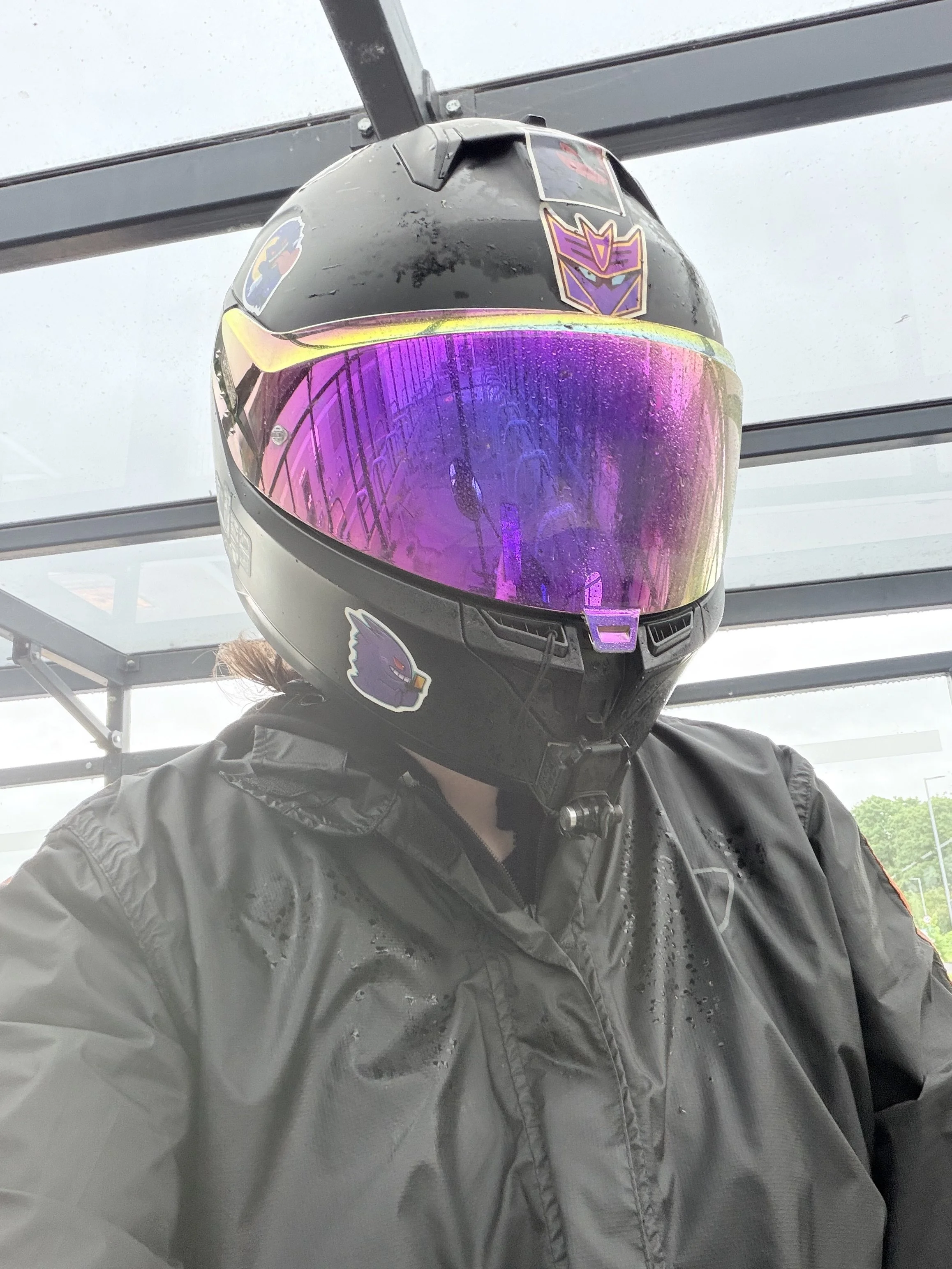 A person wearing a black motorcycle helmet with purple and yellow visor, a black jacket, standing under a glass shelter on a cloudy day.