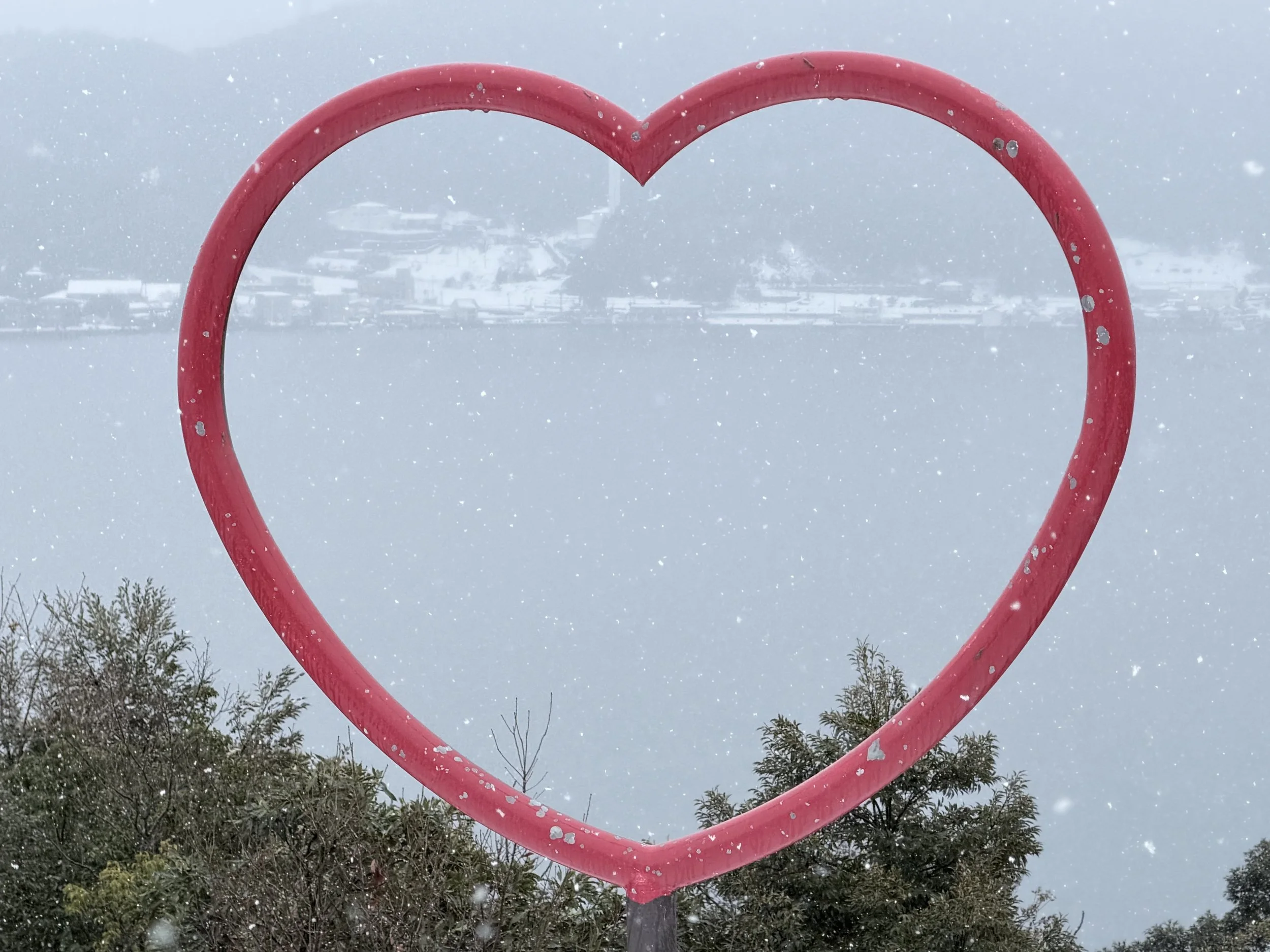 Red heart-shaped metal structure outdoors during snowfall, with trees and buildings visible in the background.