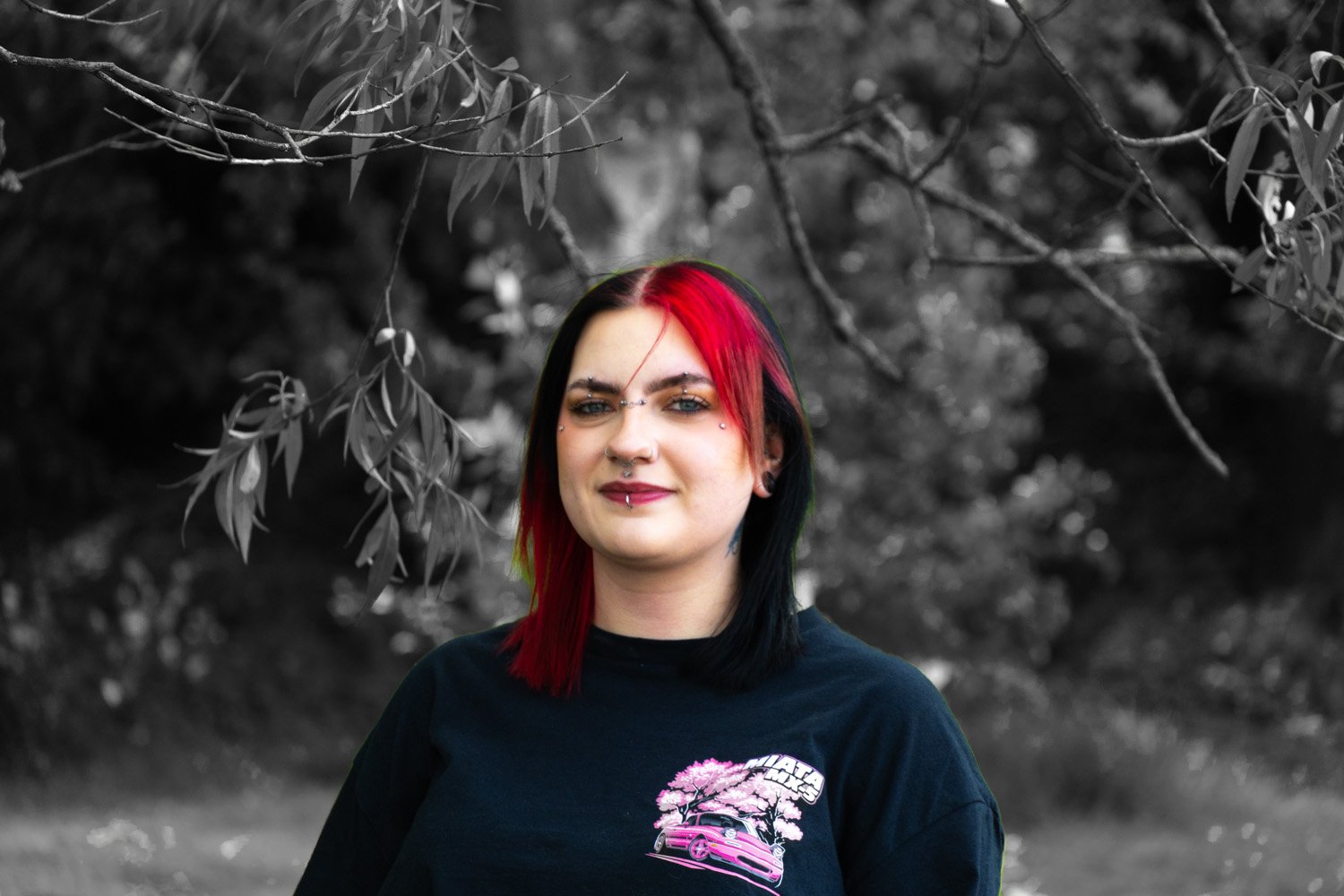 A woman with black and red hair, piercings, and tattoos outdoors, wearing a black T-shirt with a pink graphic of a car and cherry blossoms, smiling at the camera. The background is in black and white, showing trees and foliage.