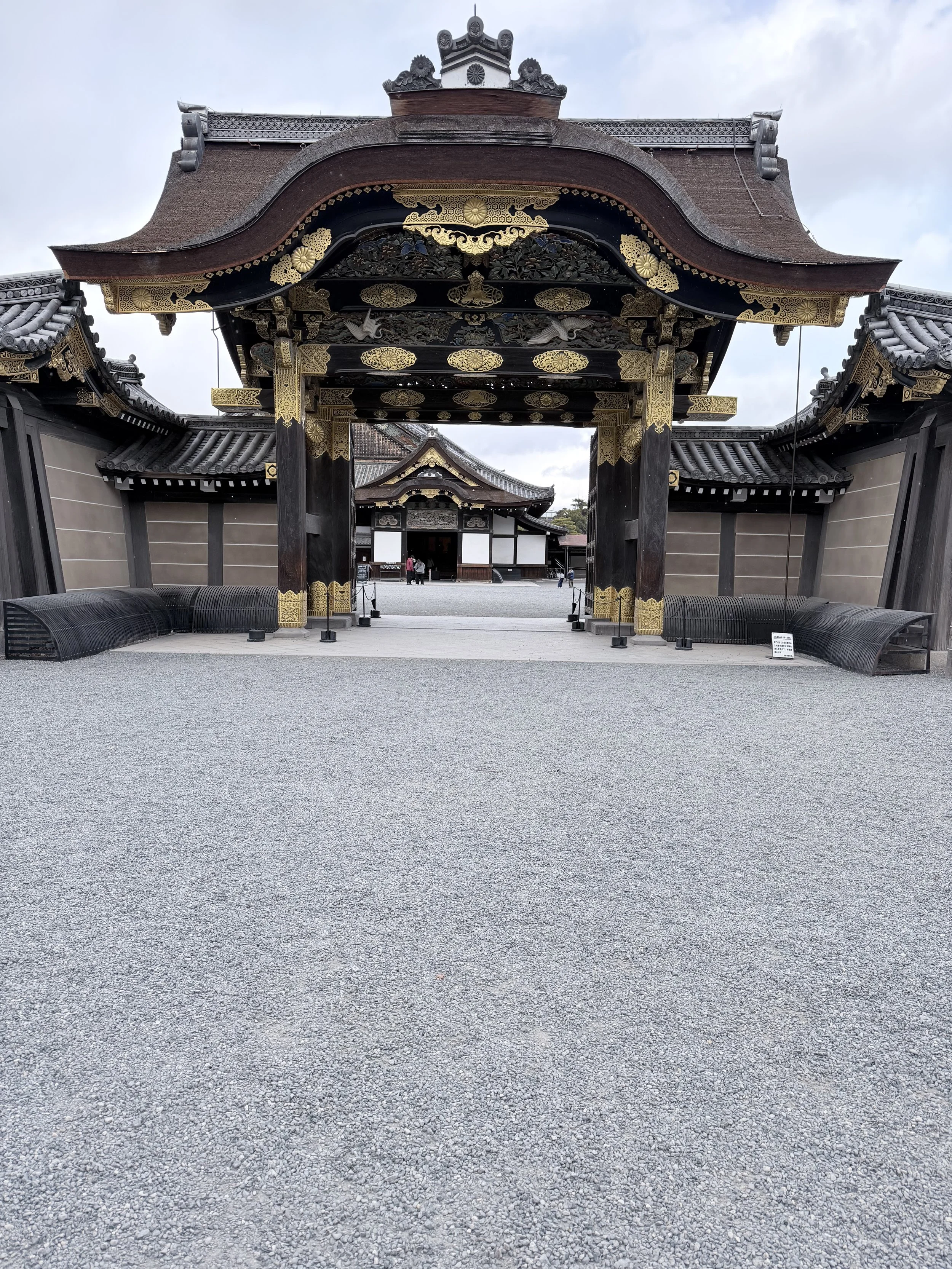 Traditional Japanese temple entrance with ornate black and gold decorations, curved roof, and gravel courtyard.