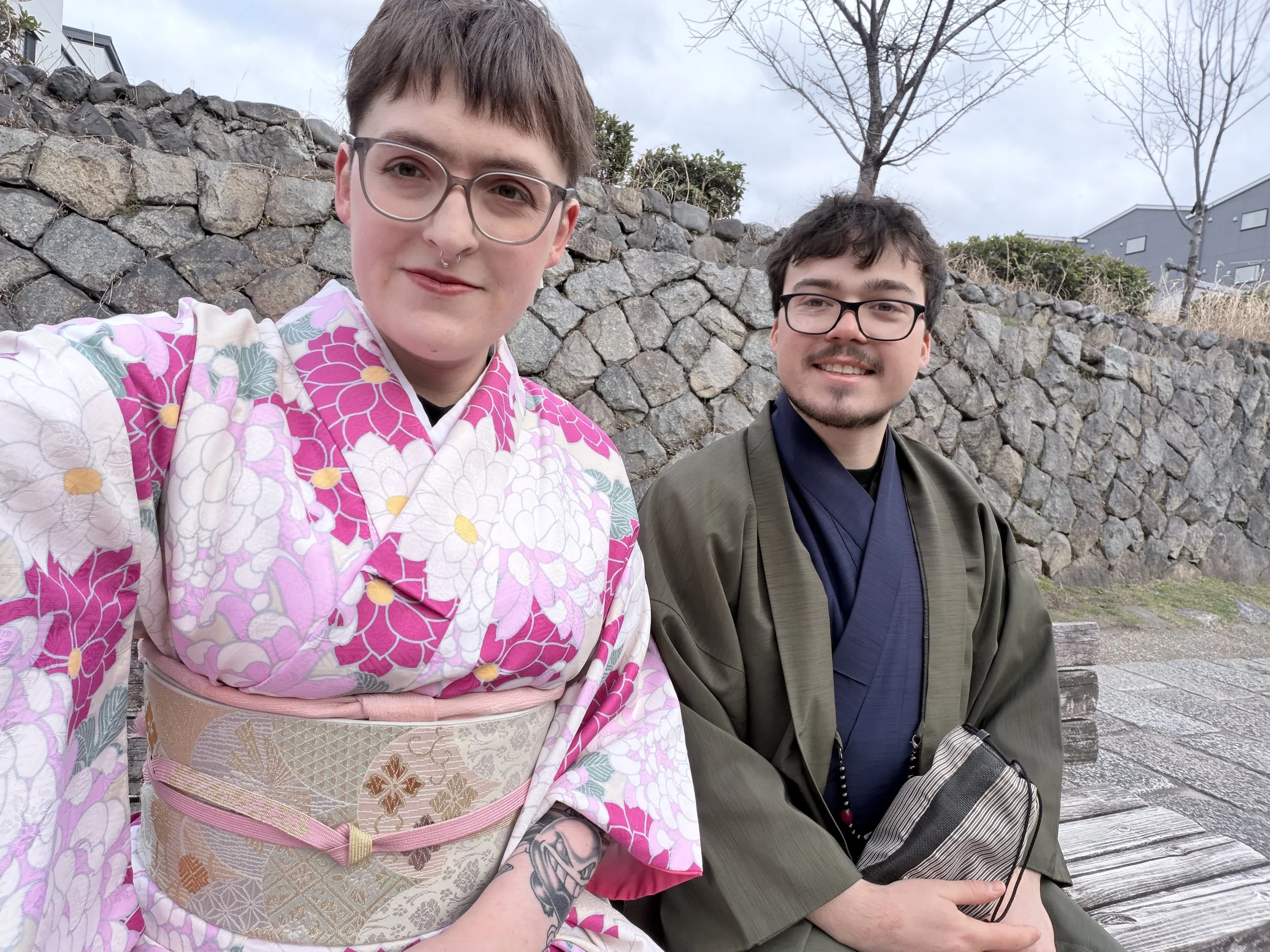Two young men sit close together outdoors in front of a stone wall, one in a kimono and the other in traditional Japanese attire, with leafless trees and buildings in the background.