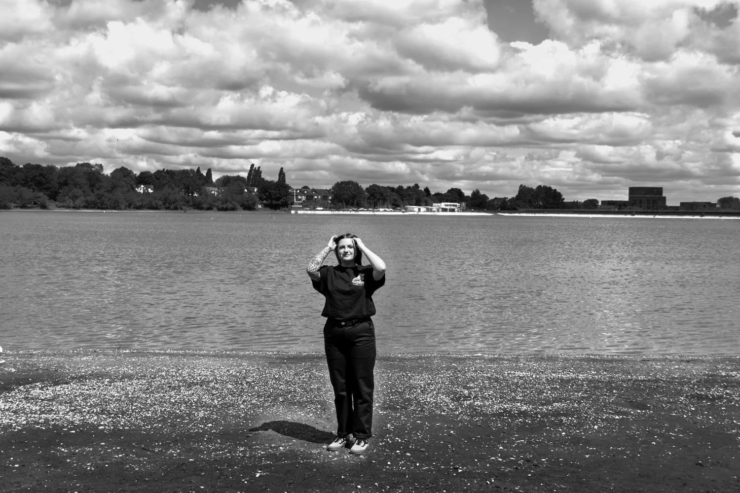 A woman standing on the shore of a lake, facing the camera, with her hands on her head, clouds in the sky, trees and buildings in the background.
