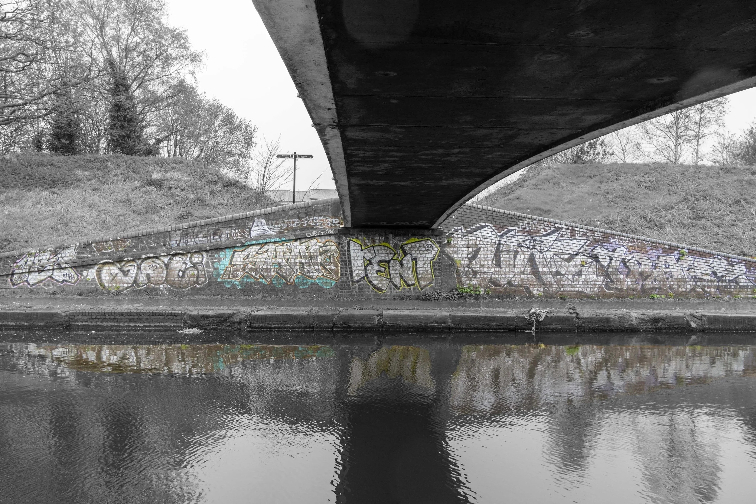 A bridge over a canal with graffiti-covered walls, trees in the background, and reflections in the water.