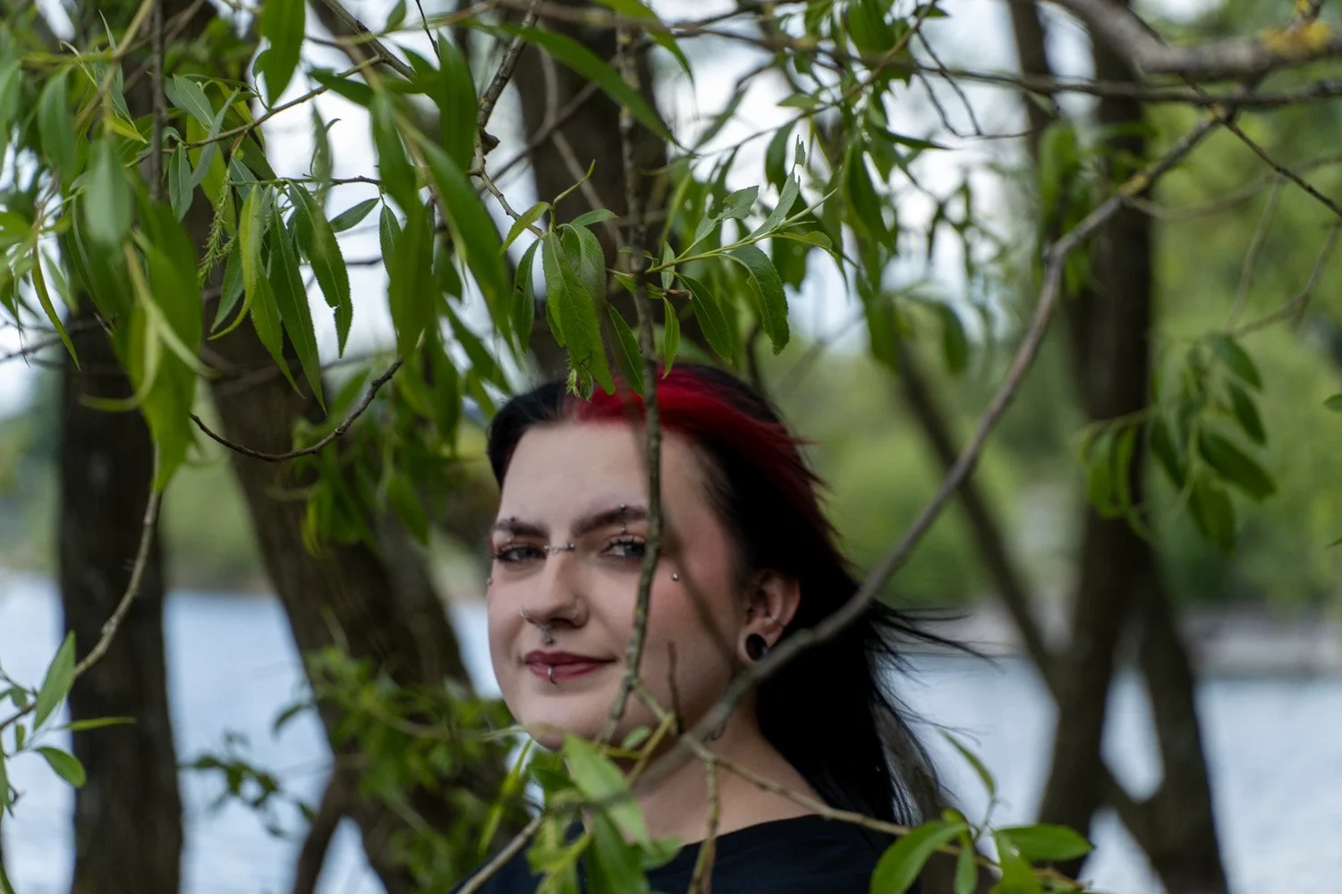 A person with dark hair and red streaks, pierced eyebrows, and piercings on their face, standing behind green leafy branches with a blurred body of water in the background.