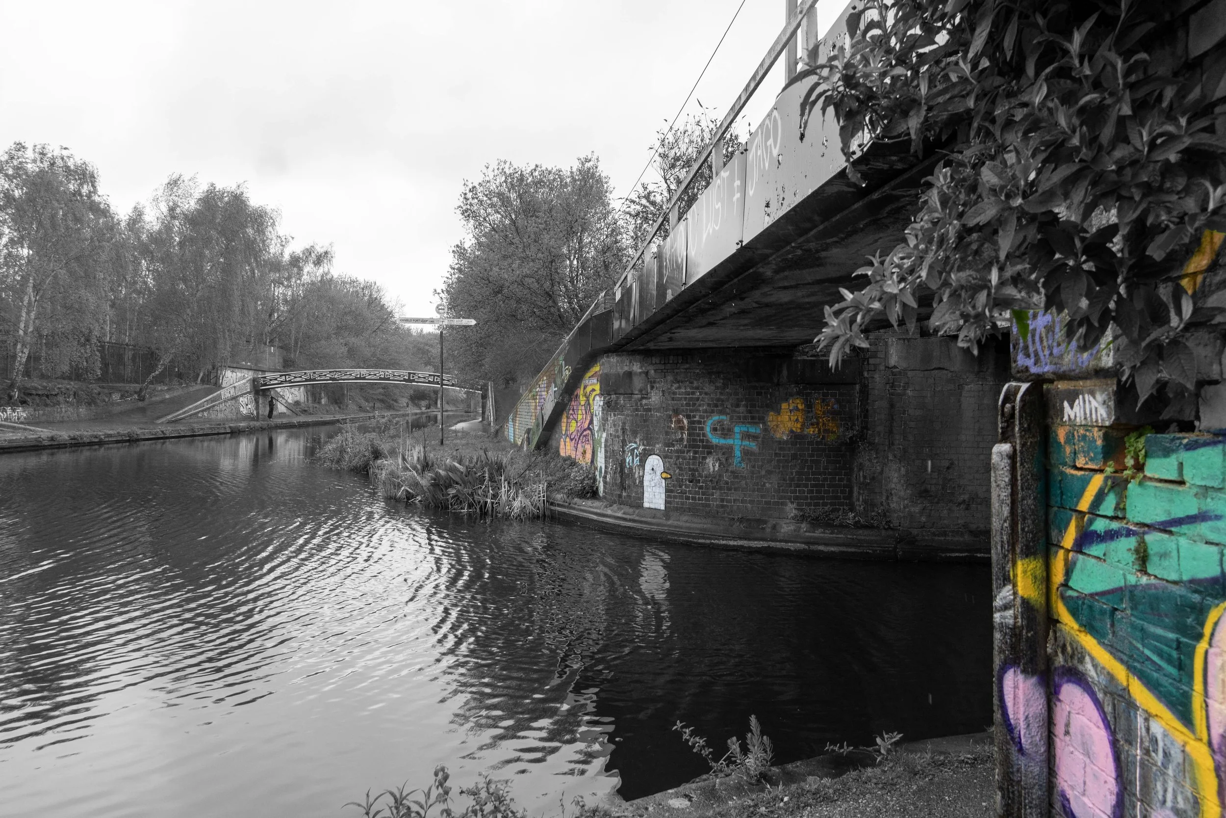 A black and white photo of a canal with a bridge and trees along the banks. A bridge with graffiti is in the foreground, and graffiti is also on the wall underneath the bridge. A person is walking along the path on the far side of the canal. The sky is overcast.