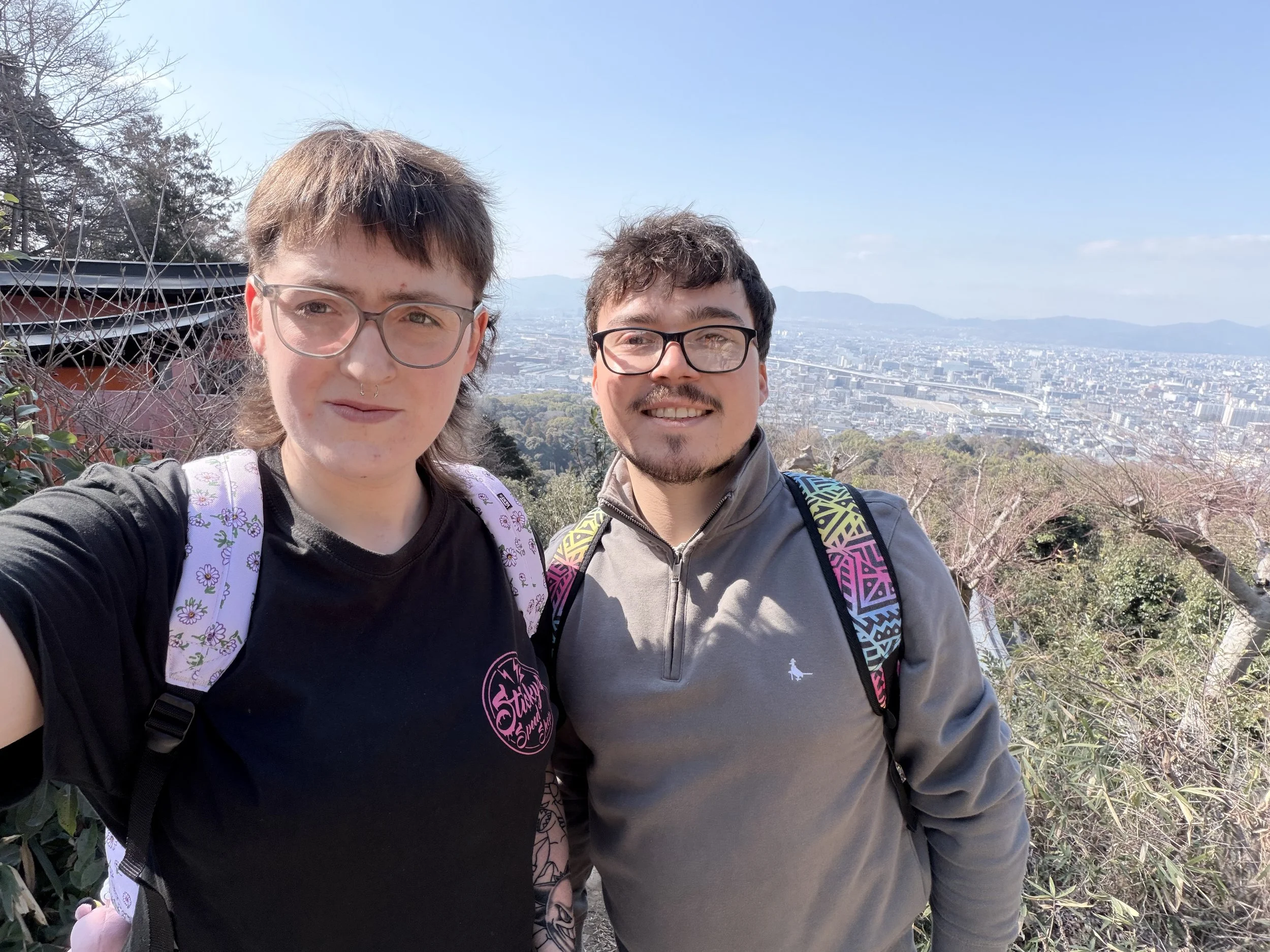 Two smiling people take a selfie outdoors with a cityscape and mountains in the background on a clear day.
