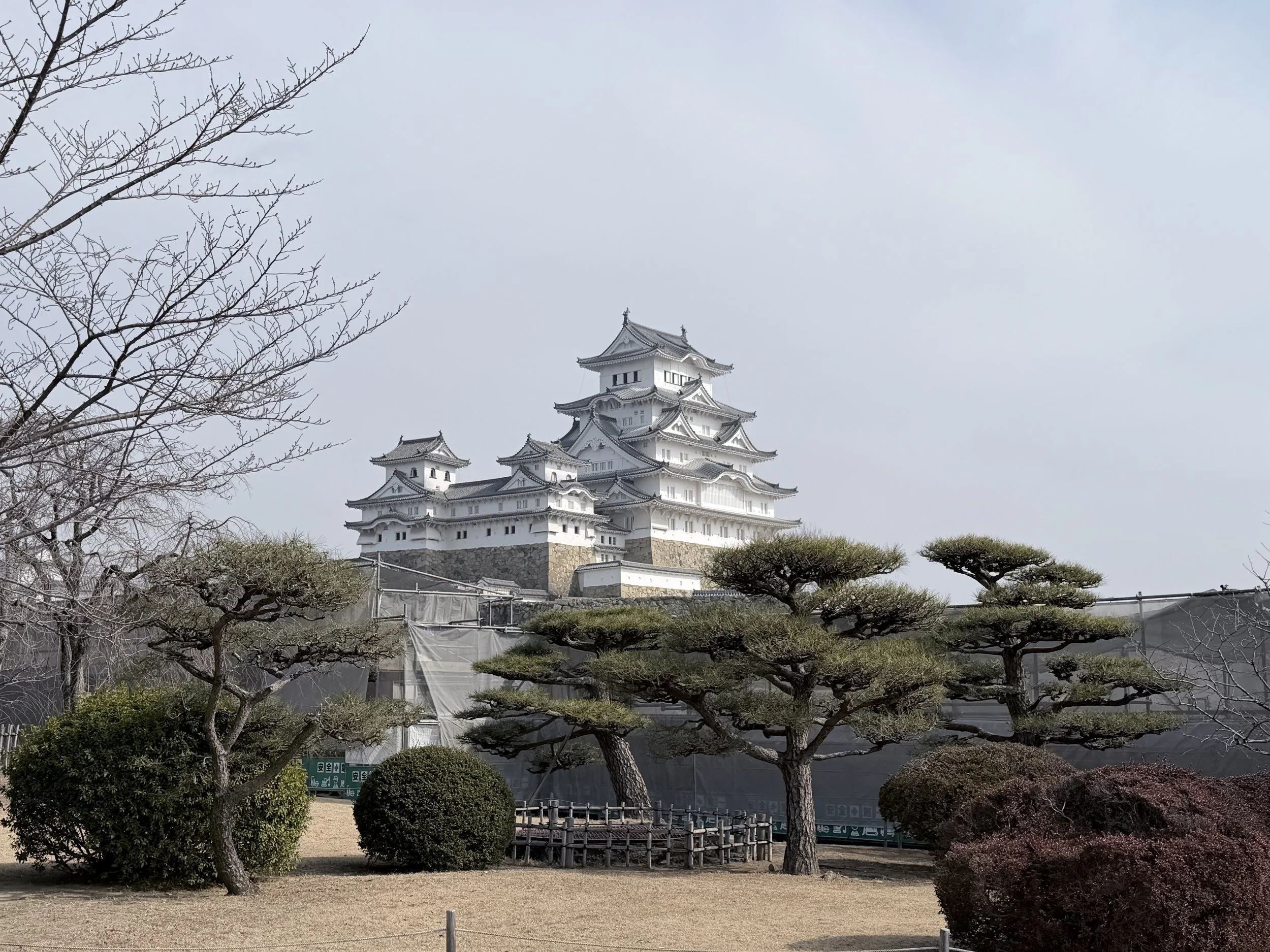 A traditional Japanese castle with white walls and multiple rooftops, surrounded by trimmed trees and bushes, under a cloudy sky.