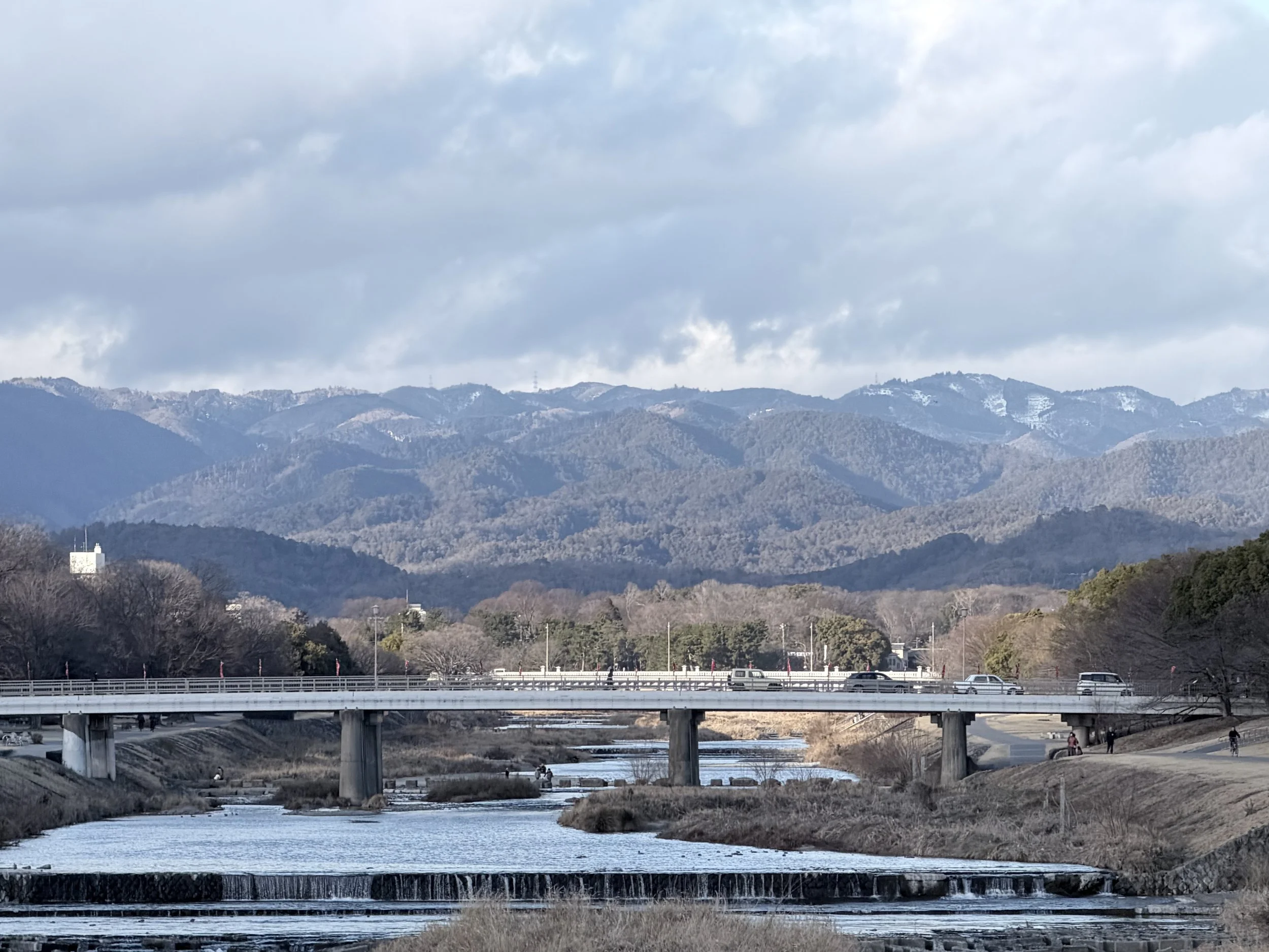A landscape scene featuring mountains with patches of snow, a river flowing under a highway bridge, and a cloudy sky overhead.