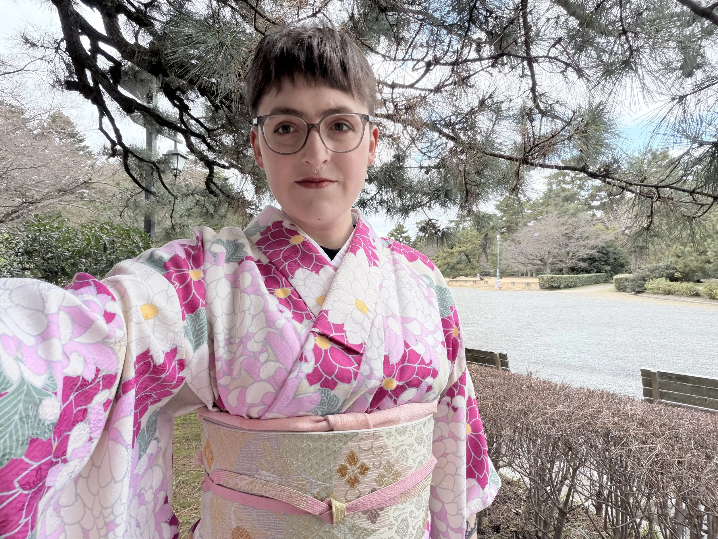 A person wearing glasses and a septum piercing taking a selfie outdoors, dressed in a pink and white floral kimono with an ornate obi, standing in front of a tree and a lake in a park.