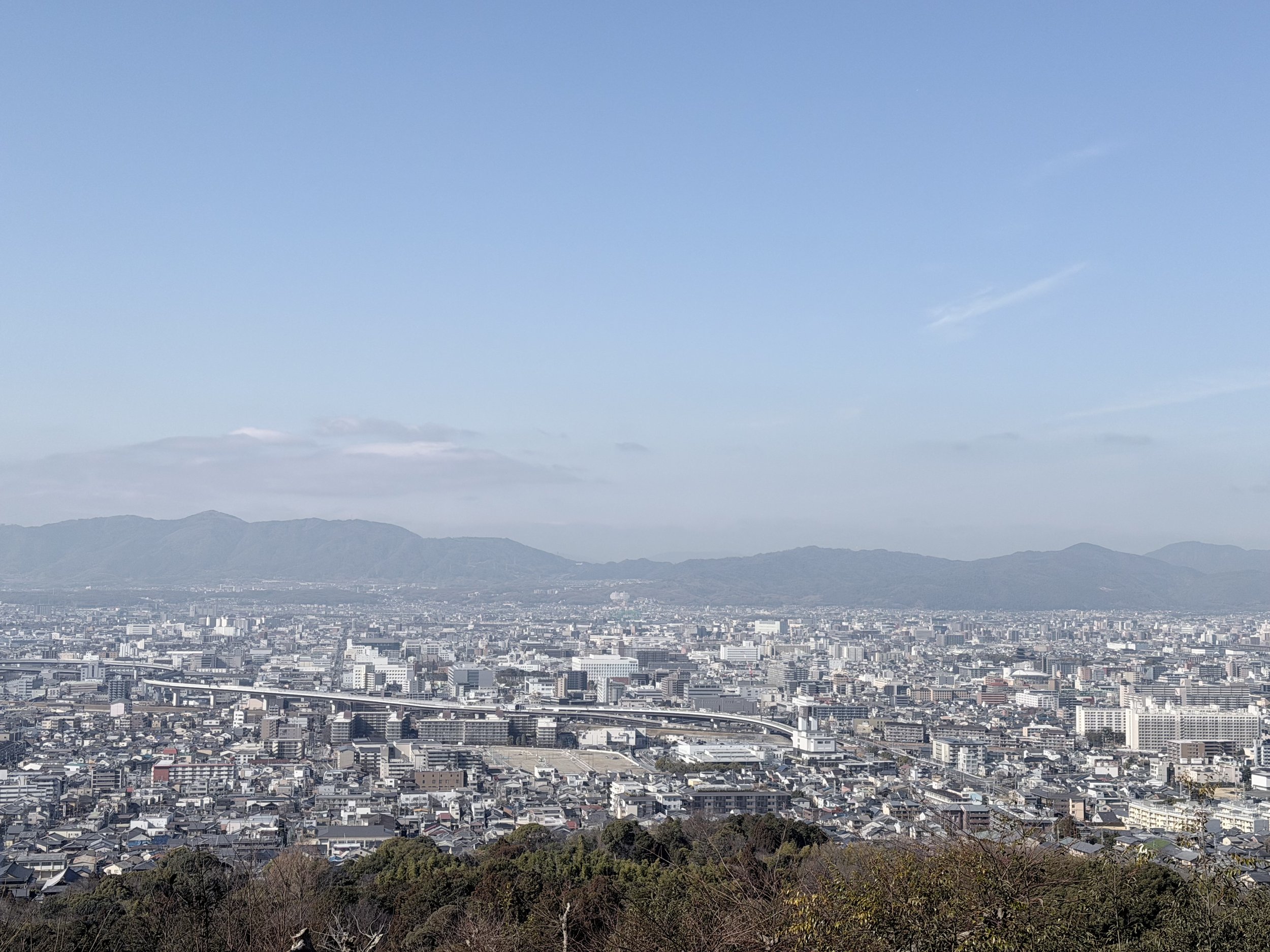 Cityscape of a densely populated urban area with numerous buildings, roads, and highways, with mountains and a clear blue sky in the background.