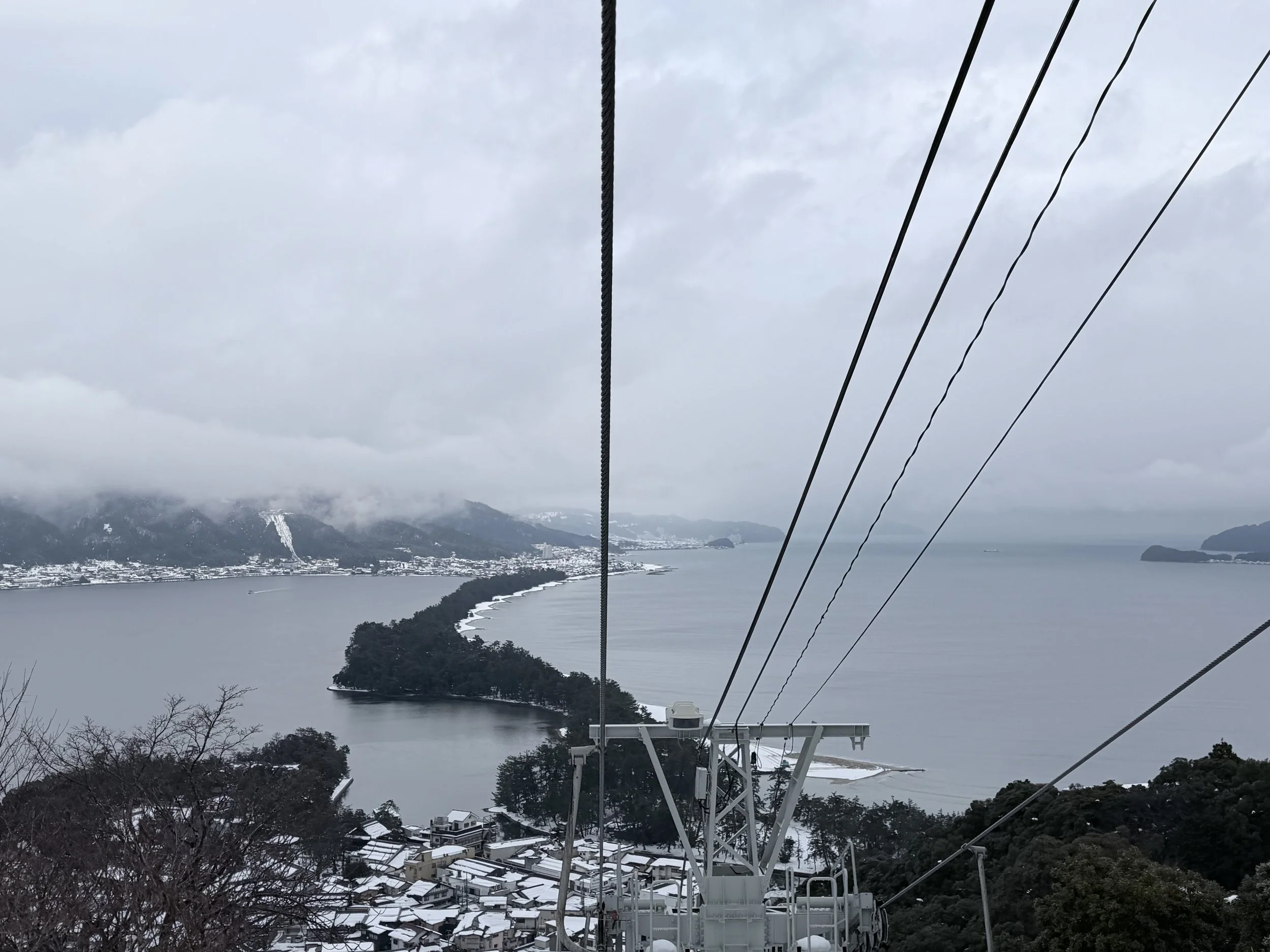 View from a cable car over a snowy landscape with mountains, water, and buildings in the distance, cloudy sky.