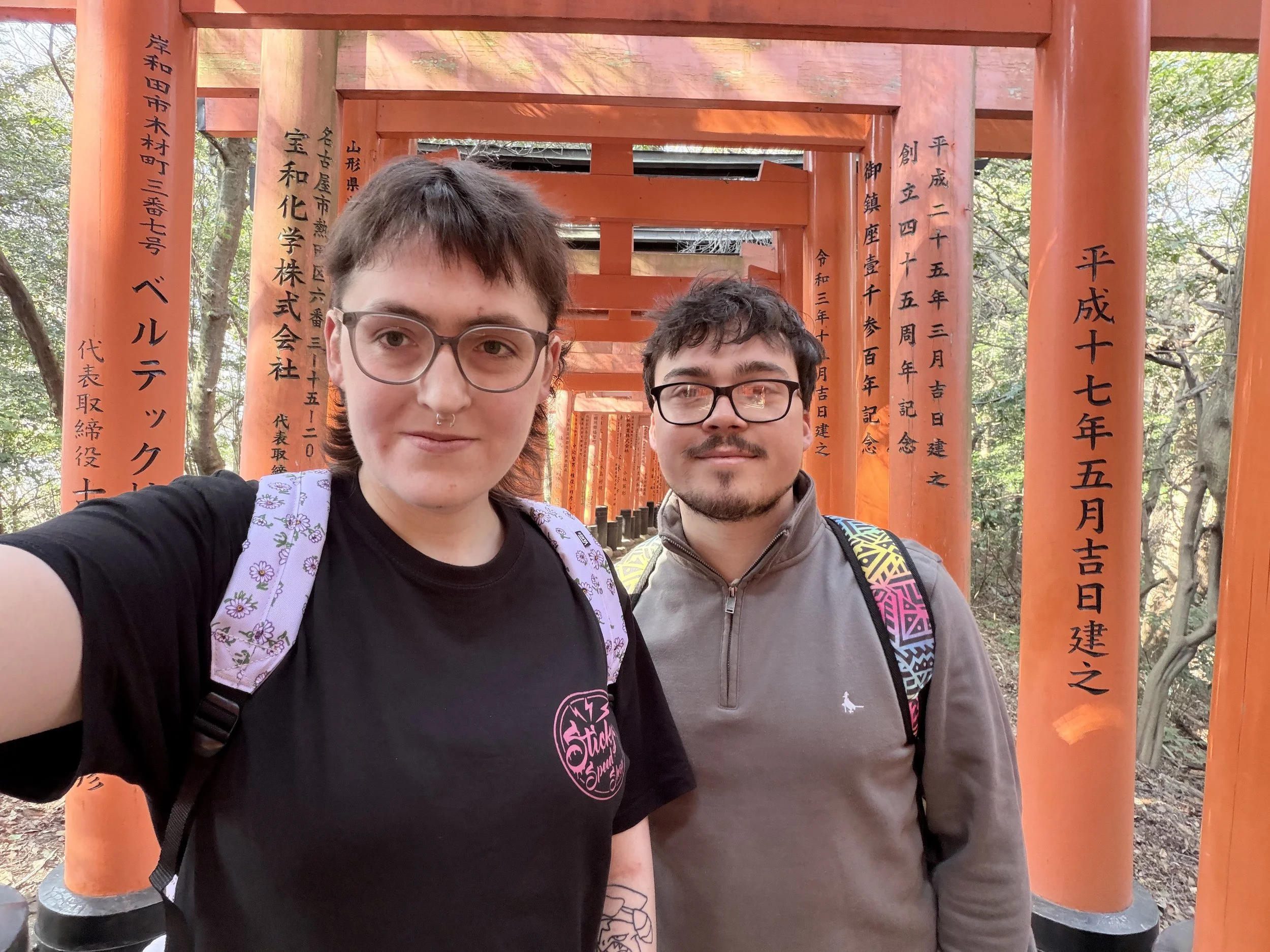 Two young people, one with short curly dark hair and glasses, the other with short light brown hair and glasses, standing in front of a series of orange torii gates at a Japanese shrine or temple, surrounded by trees.