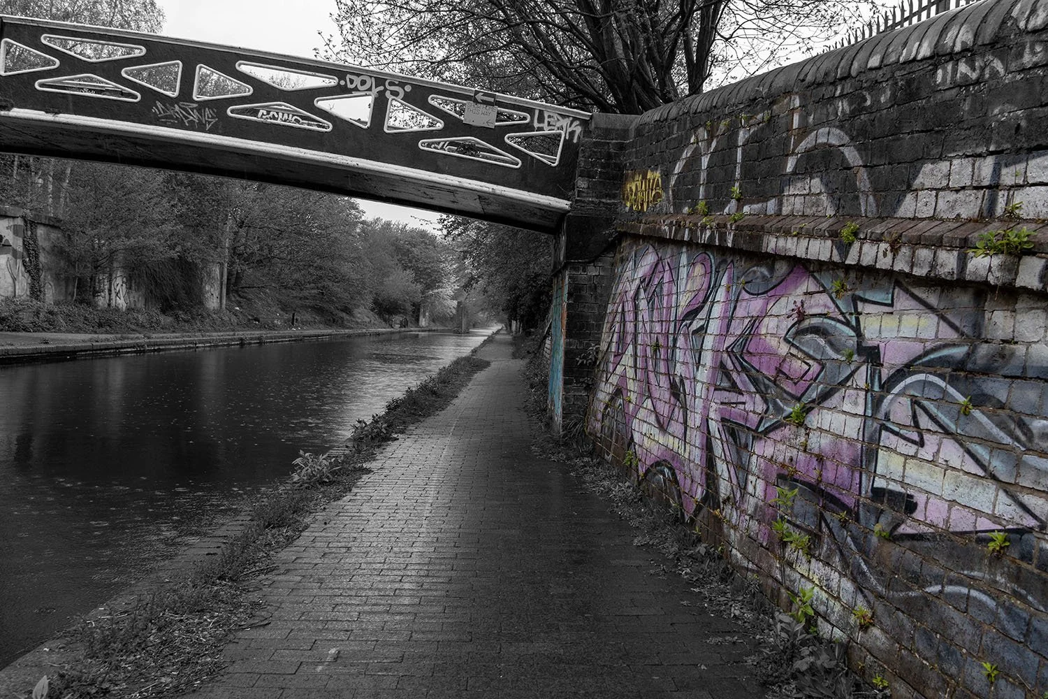 A black and white photo of a canal with a paved walkway on the right side, bordered by a graffiti-covered brick wall. A bridge with graffiti and cut-out patterns crosses over the canal in the background, with trees lining the canal on the left.