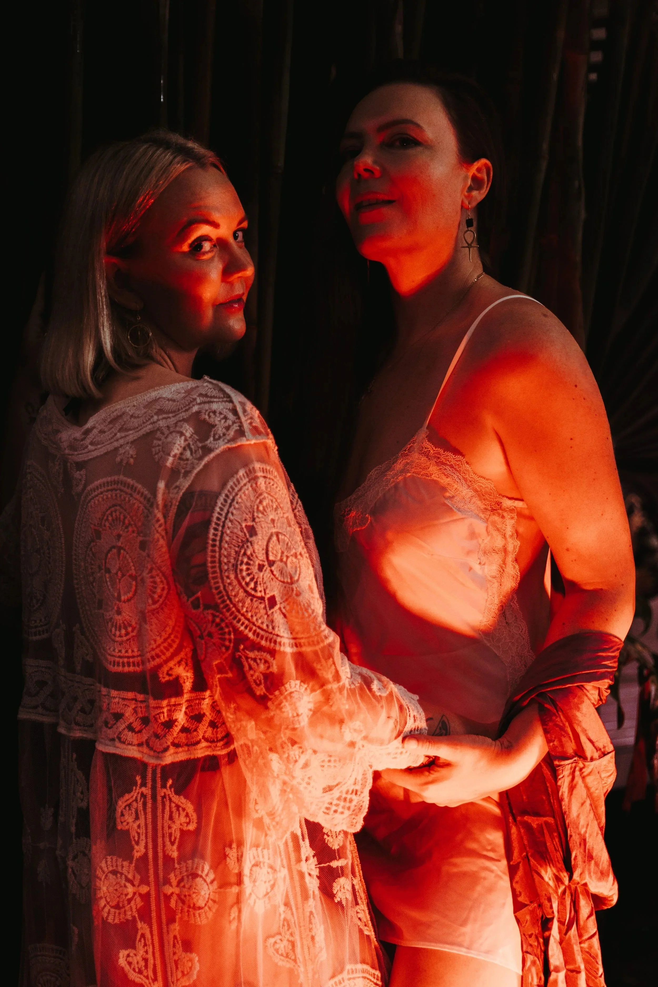 Two women in lace and satin lingerie, one with acrylic nails, earrings and a bracelet, are posing in a dark environment with red lighting, possibly at a party or a photoshoot.