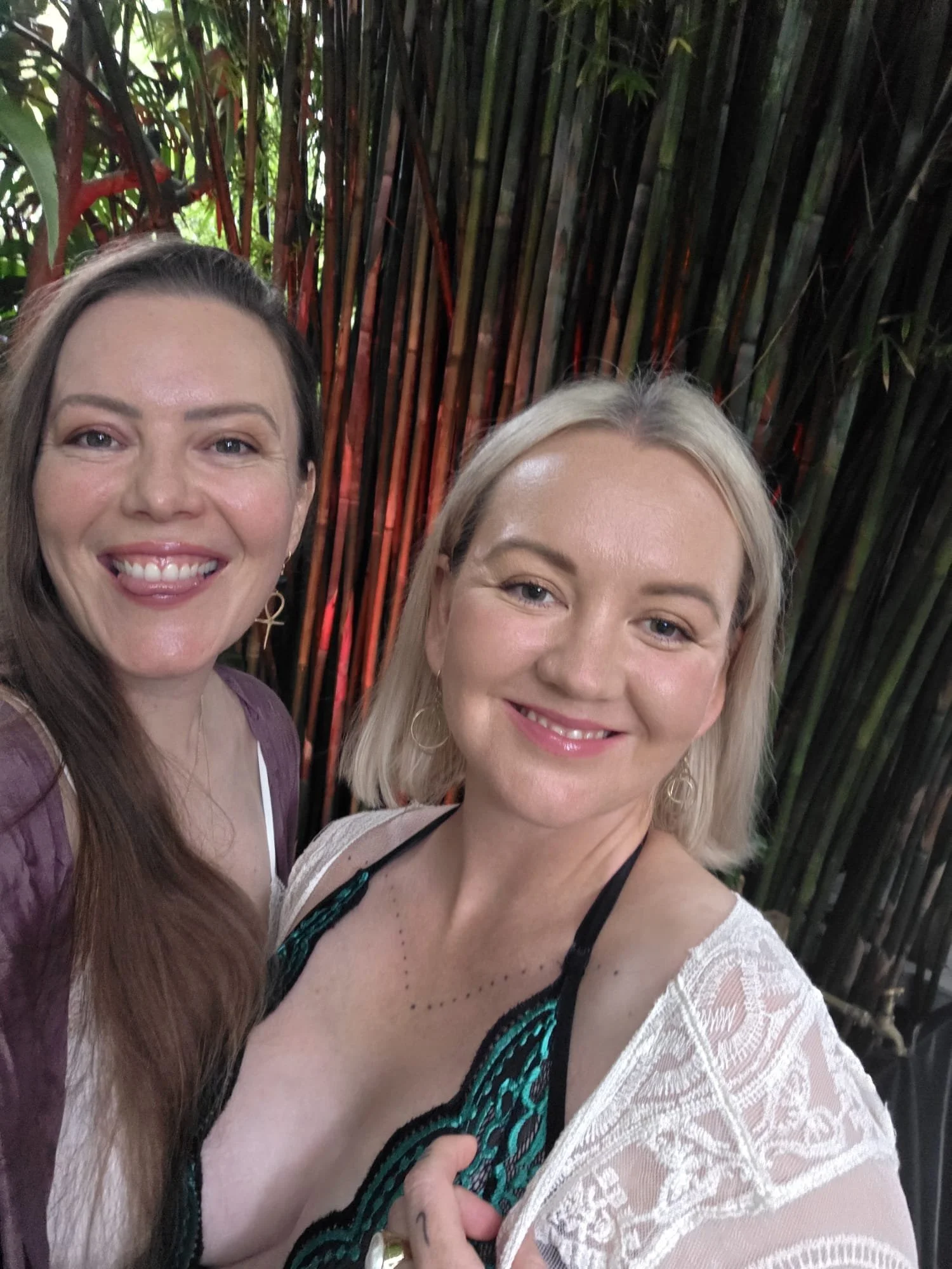 Two women are taking a selfie in front of green bamboo plants, smiling at the camera.