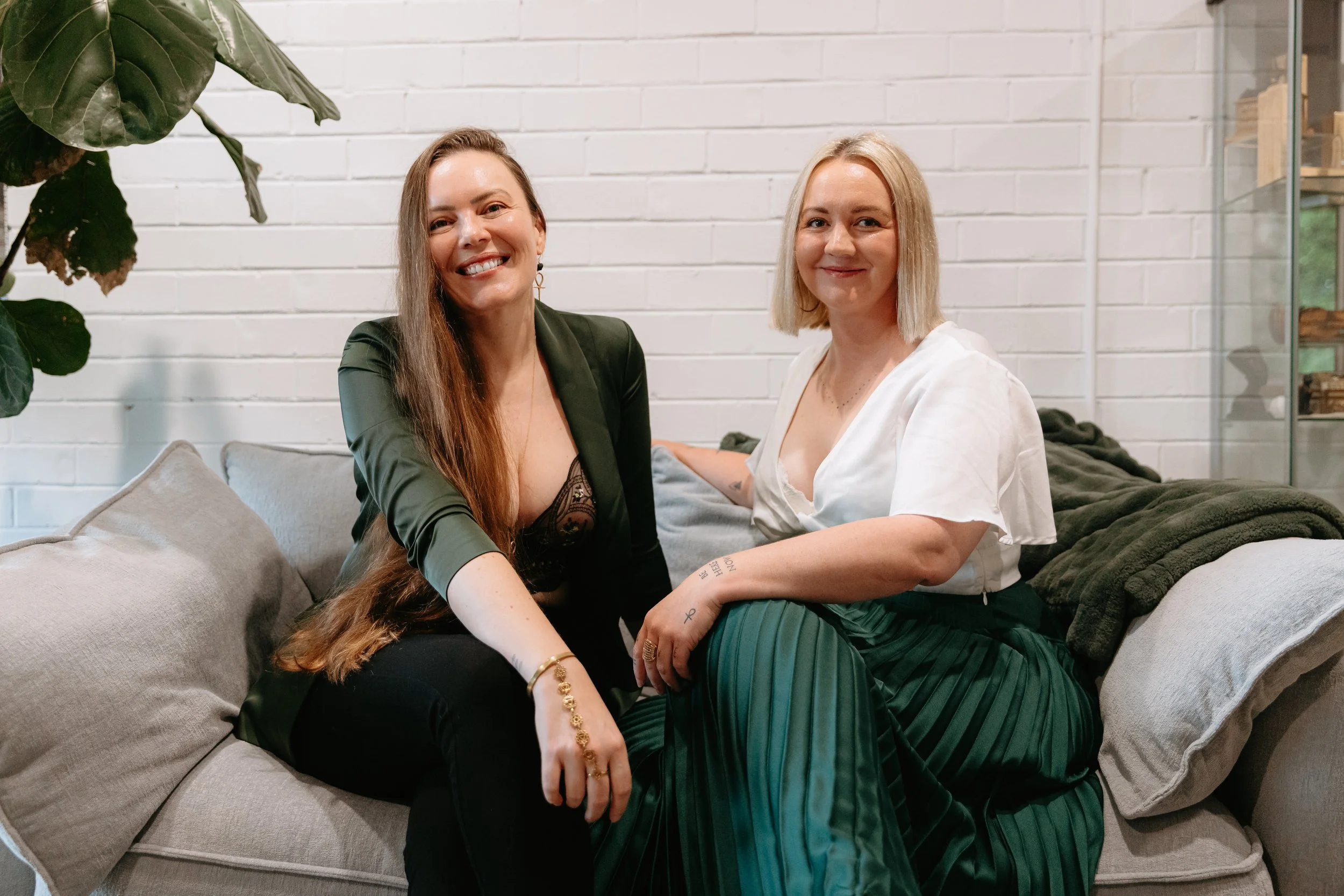 Two women sitting on a gray couch in front of a white brick wall, smiling at the camera. One woman has long brown hair and wears a black blazer, the other has short blonde hair and wears a white blouse. Large green plant leaves are partially visible on the left.