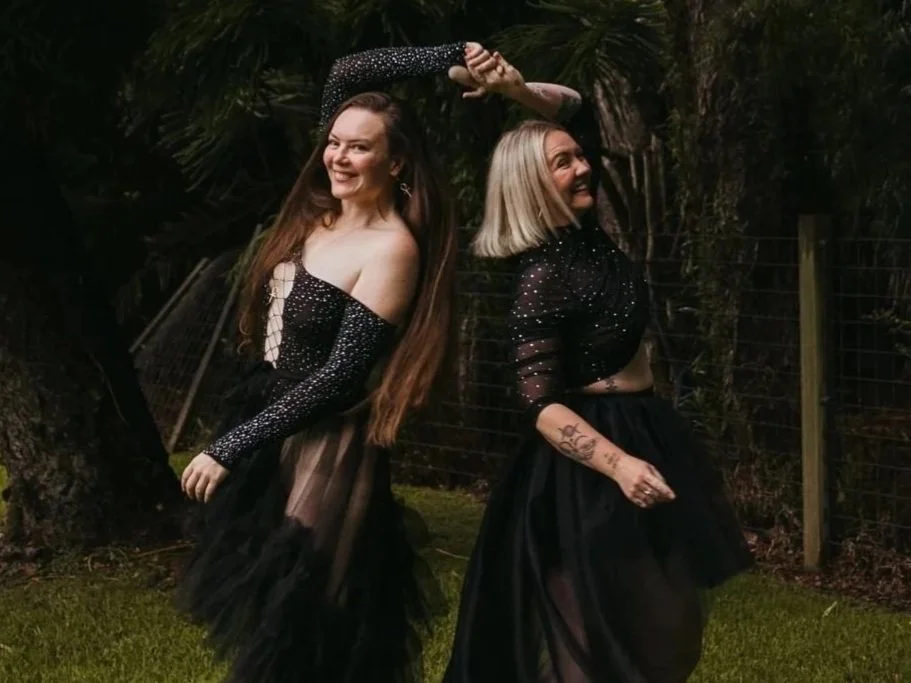 Two women dressed in black, semi-sheer, sparkly skirts and tops, standing outdoors near a tree and a fence, smiling and posing Playfully.