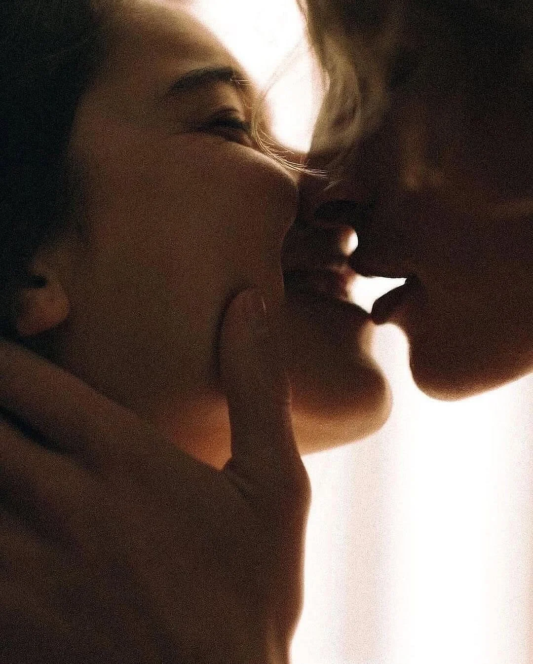 Close-up of two people about to kiss, with one person's finger gently touching the other's chin. Silhouettes created by soft backlight, emphasizing their facial features and intimate moment.