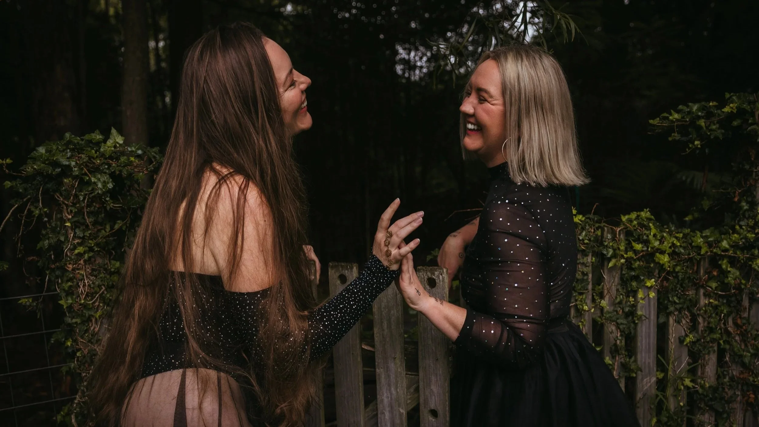 Two women with long hair are smiling and touching hands in an outdoor garden setting at dusk, surrounded by greenery and a wooden fence.