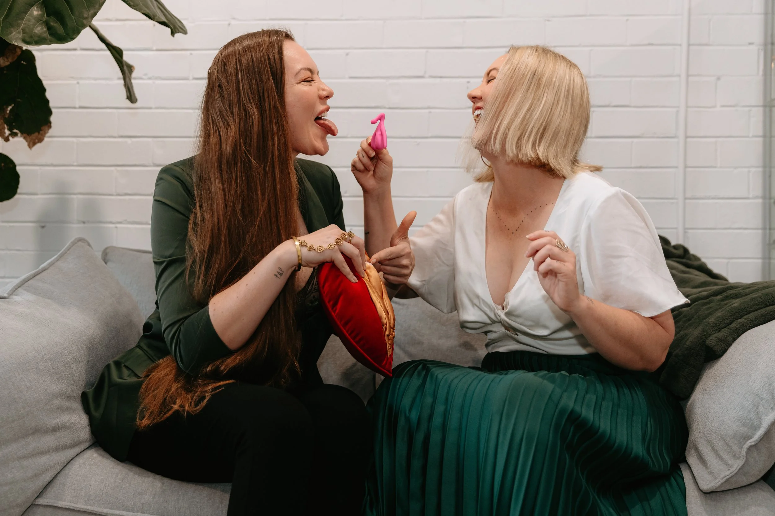 Two women sitting on a gray couch against a white brick wall, engaging playfully with a pink toy, smiling and laughing with their tongues out.