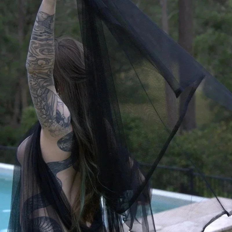 Woman with long hair and tattoos holding a black sheer fabric near her face outdoors near a pool.