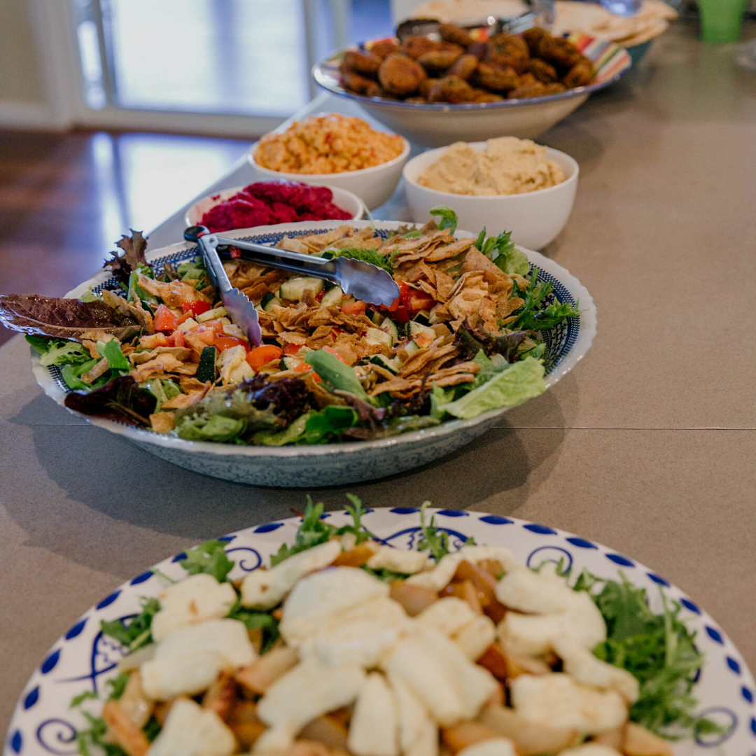 A spread of various dishes on a kitchen counter, including a large salad with greens, shredded chicken, and vegetables, along with bowls of hummus, mashed potatoes, beet salad, and a plate of fried chicken or chicken wings.