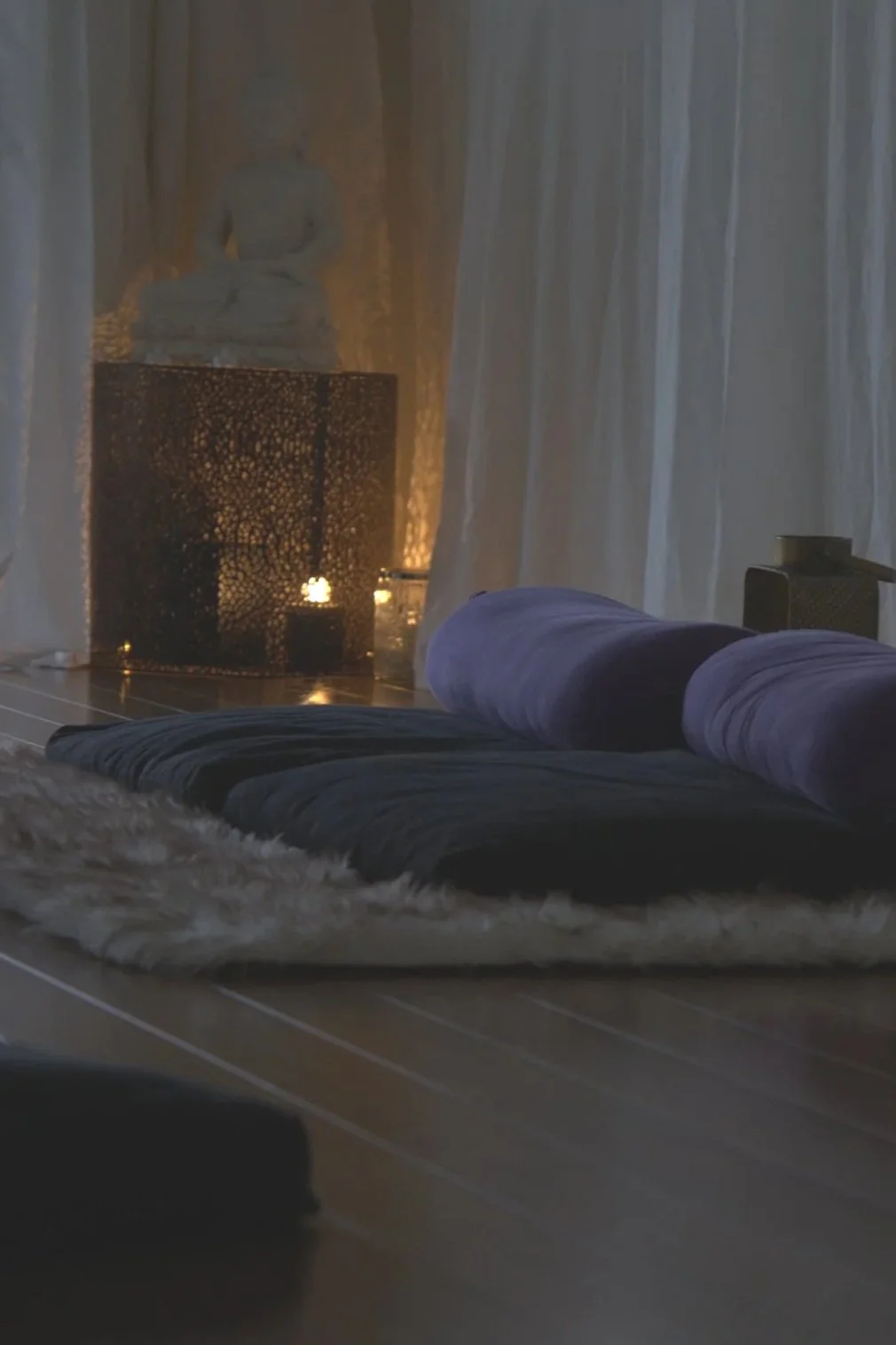 Perspective of a cozy room with a bed with pillows, sheer curtains, a lit candle holder, and a Buddha statue in the background, creating a relaxing ambiance.