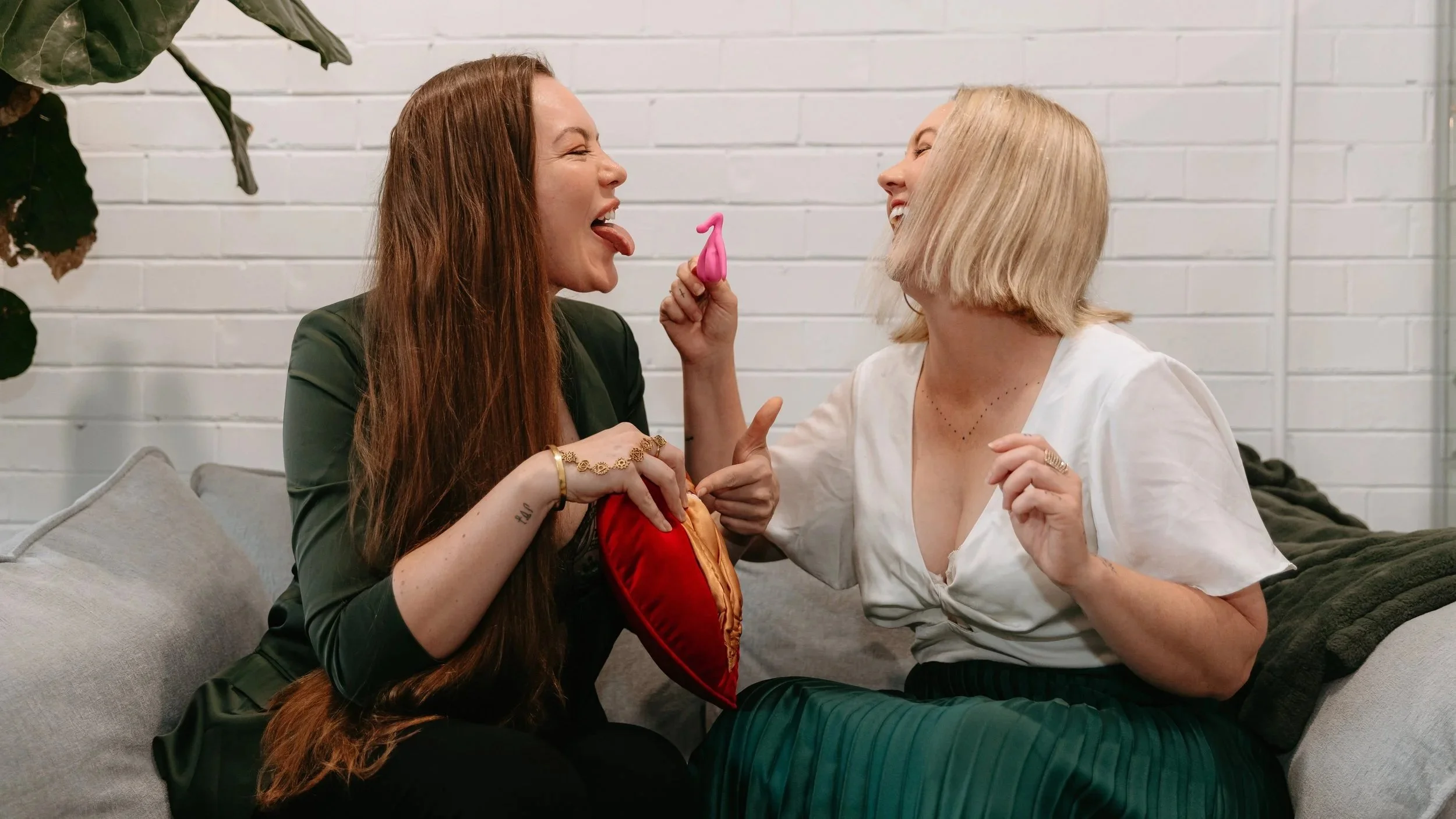 Two women sitting on a couch, playfully tugging at a pink toy, laughing and sticking their tongues out at each other, with a white brick wall in the background.