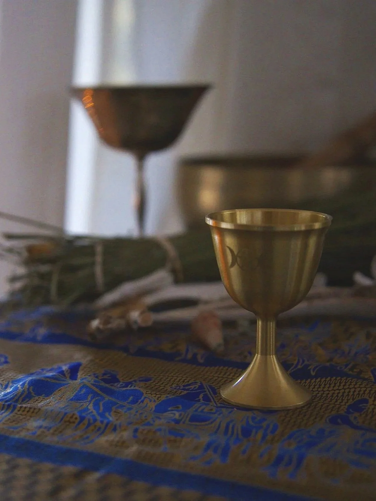 A close-up of a gold chalice on a decorative cloth with blue and gold patterns. In the background, there are other metallic objects and glasses, slightly out of focus.