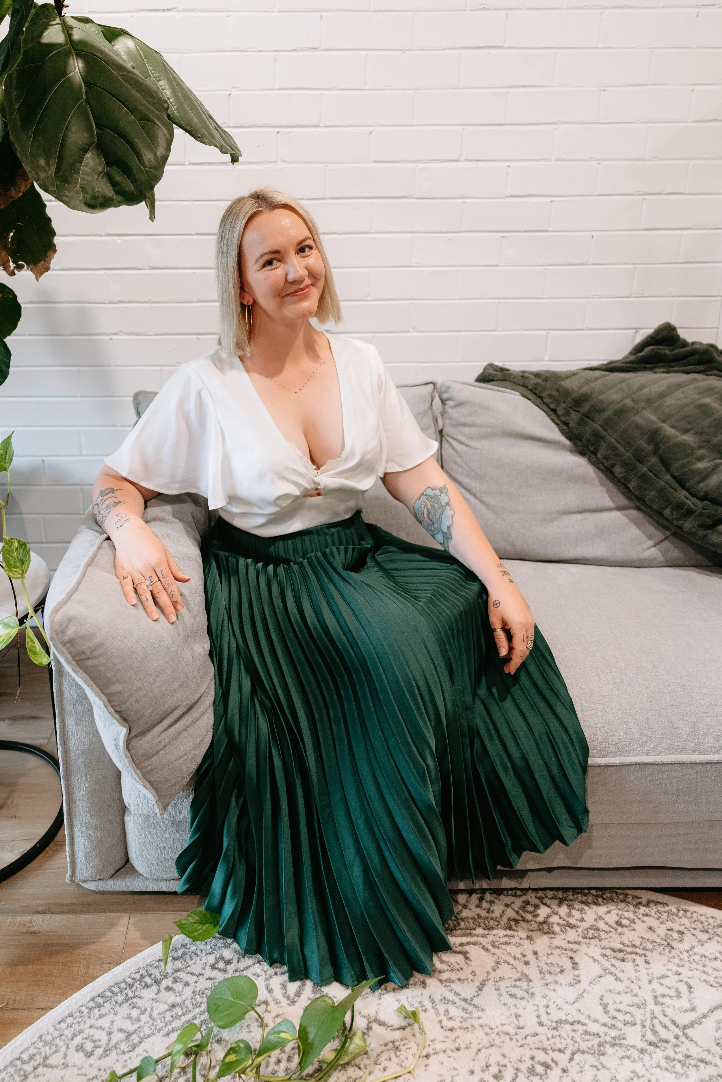 A woman with blonde hair, wearing a white blouse and a dark green pleated skirt, sitting on a beige armchair with a light gray pillow and a green blanket. There are green plants to her left, a white brick wall behind her, and a patterned rug on the wooden floor.
