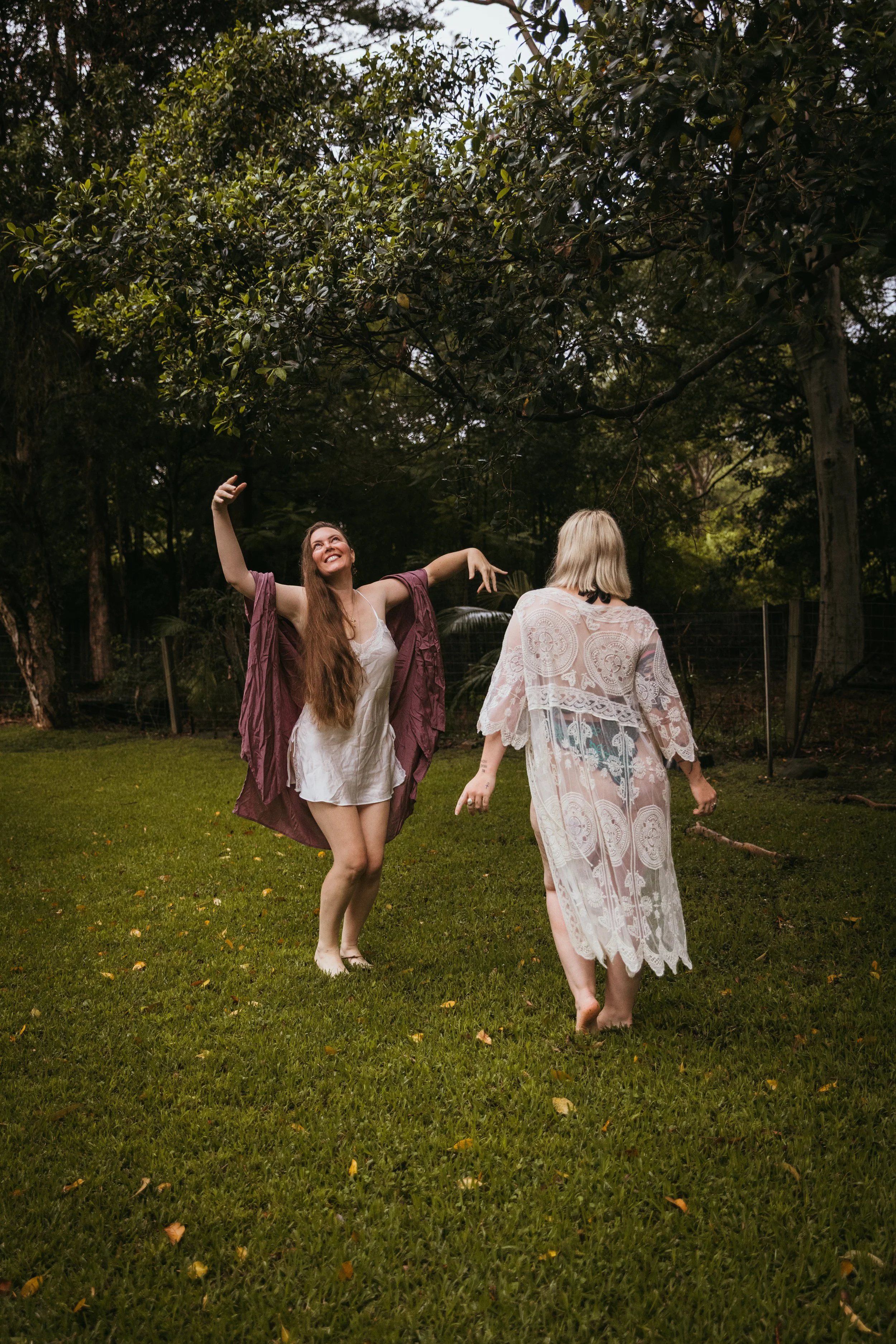 Two women in a grassy backyard with trees, one with long red hair wearing a white dress and purple cover-up, dancing and smiling, and the other with blonde hair in a patterned dress walking away.