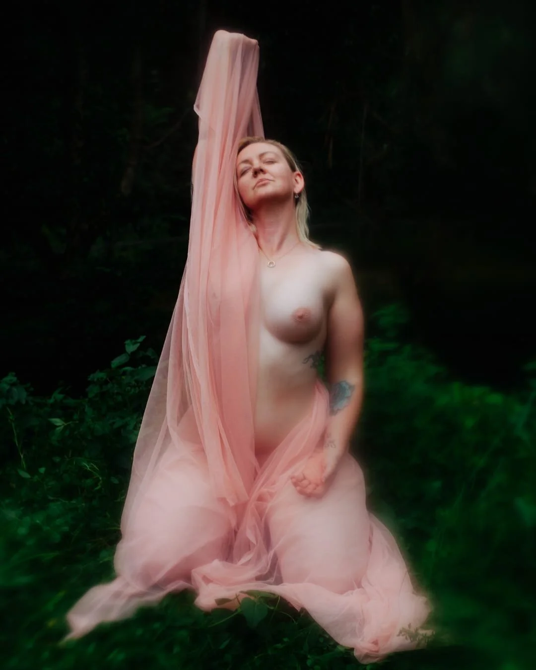 A woman with blonde hair, sitting nude in a lush green forest, holding a flowing pink fabric that drapes over her body and extends above her head.