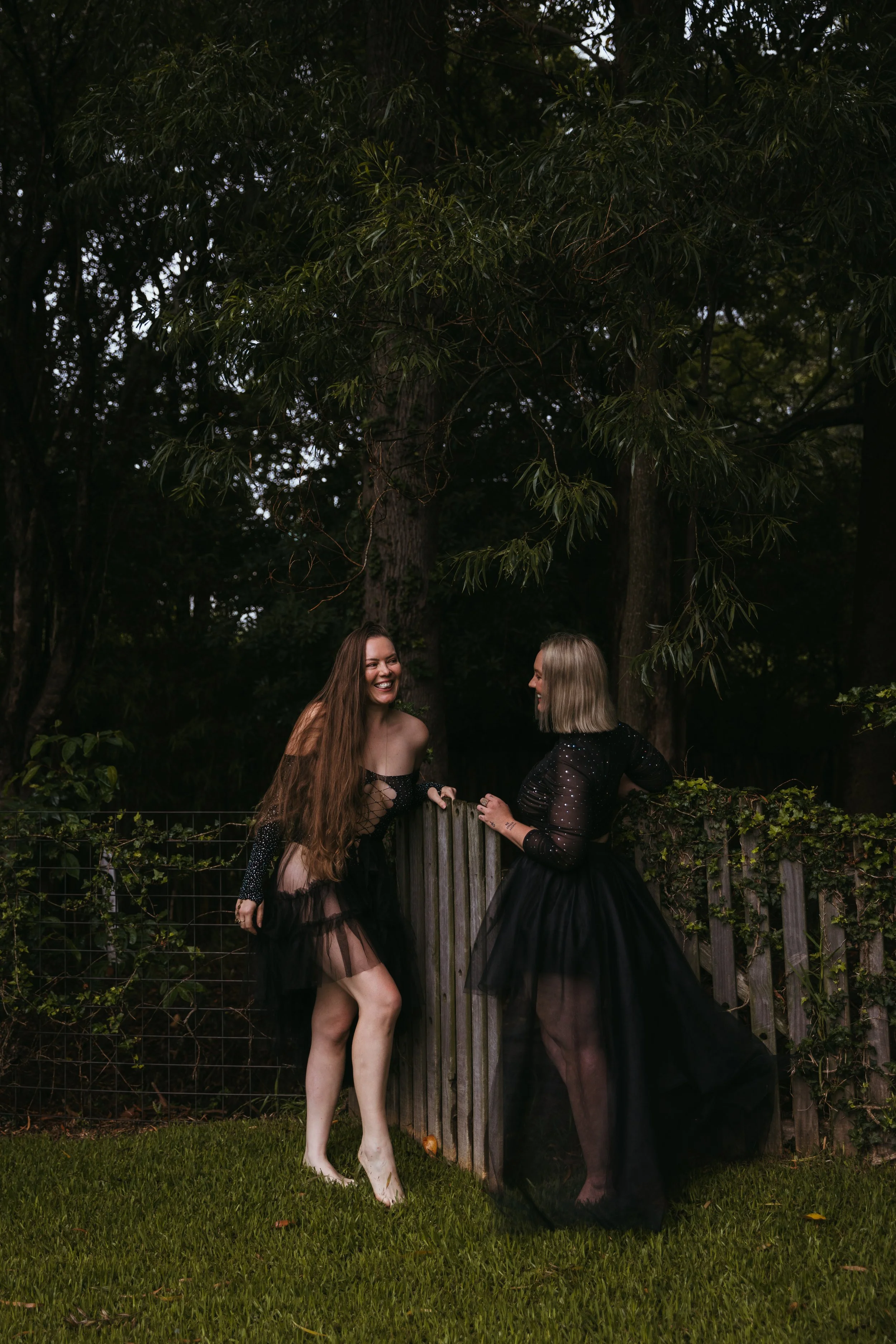Two women in black sheer dresses with long skirts, standing outdoors on grass, smiling and talking to each other, with a wooden fence and green trees in the background.