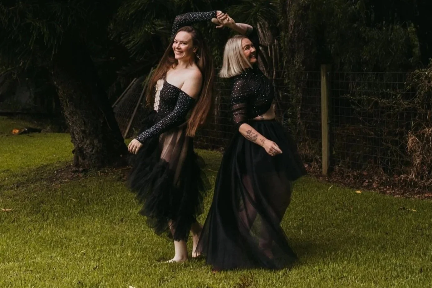 Two women with long and short blonde hair respectively, wearing black, sparkly, mesh long dresses with sheer skirts, standing in a grassy area outdoors at night. They are smiling and posing creatively with one woman having her arm extended above her head and the other holding her dress. A tree and a wire fence are visible in the background.