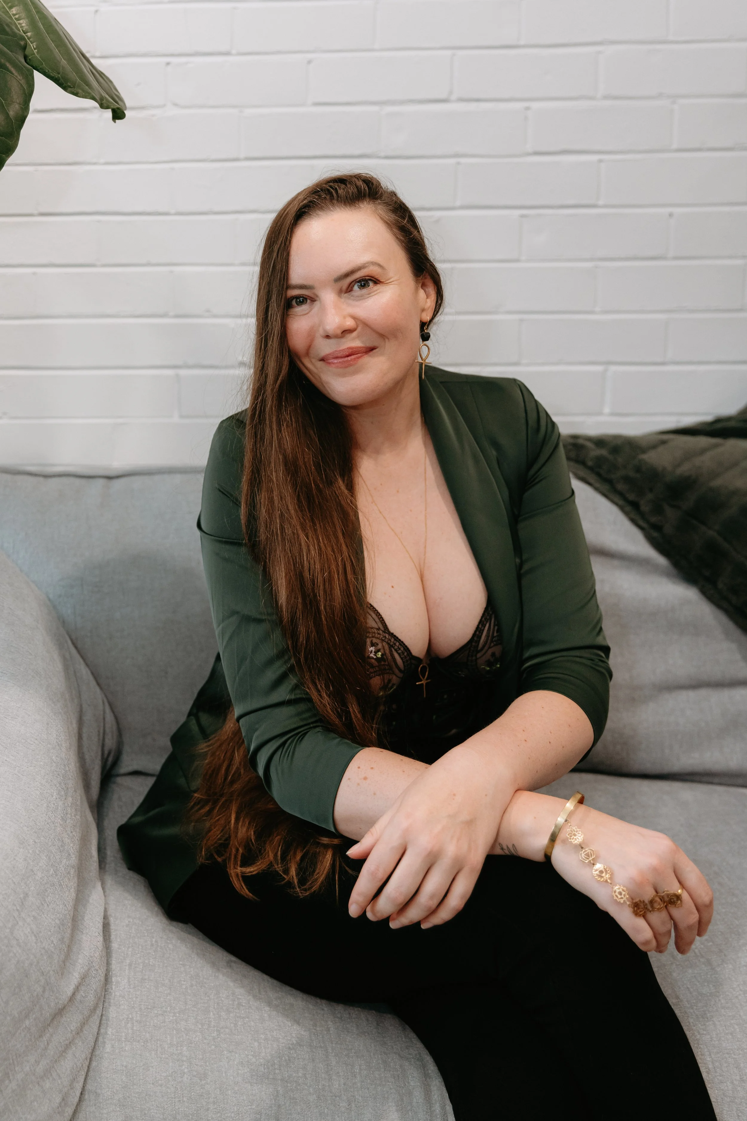 A woman with long brown hair, wearing a black lace top and an olive green blazer, sitting on a light gray sofa with a white brick wall in the background.