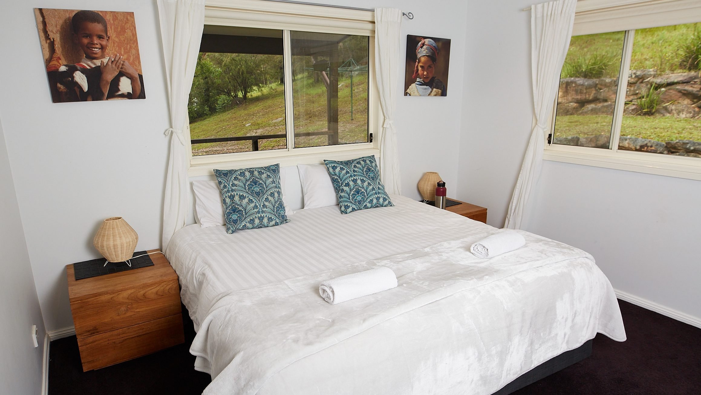A bedroom with a large bed dressed in white linens, two rolled white towels at the foot, and decorative blue pillows. Two wooden nightstands with wicker lamps sit beside the bed. There are two large windows with white curtains, showing a grassy outdoor scene. Two framed photographs of children hang on the white walls.