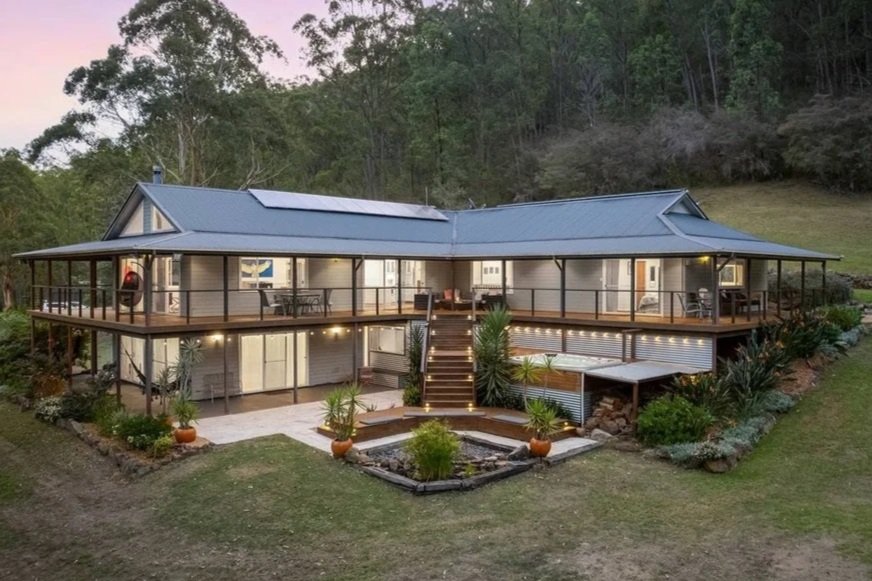 Large two-story house with a wrap-around deck, surrounded by greenery and trees, illuminated at dusk.